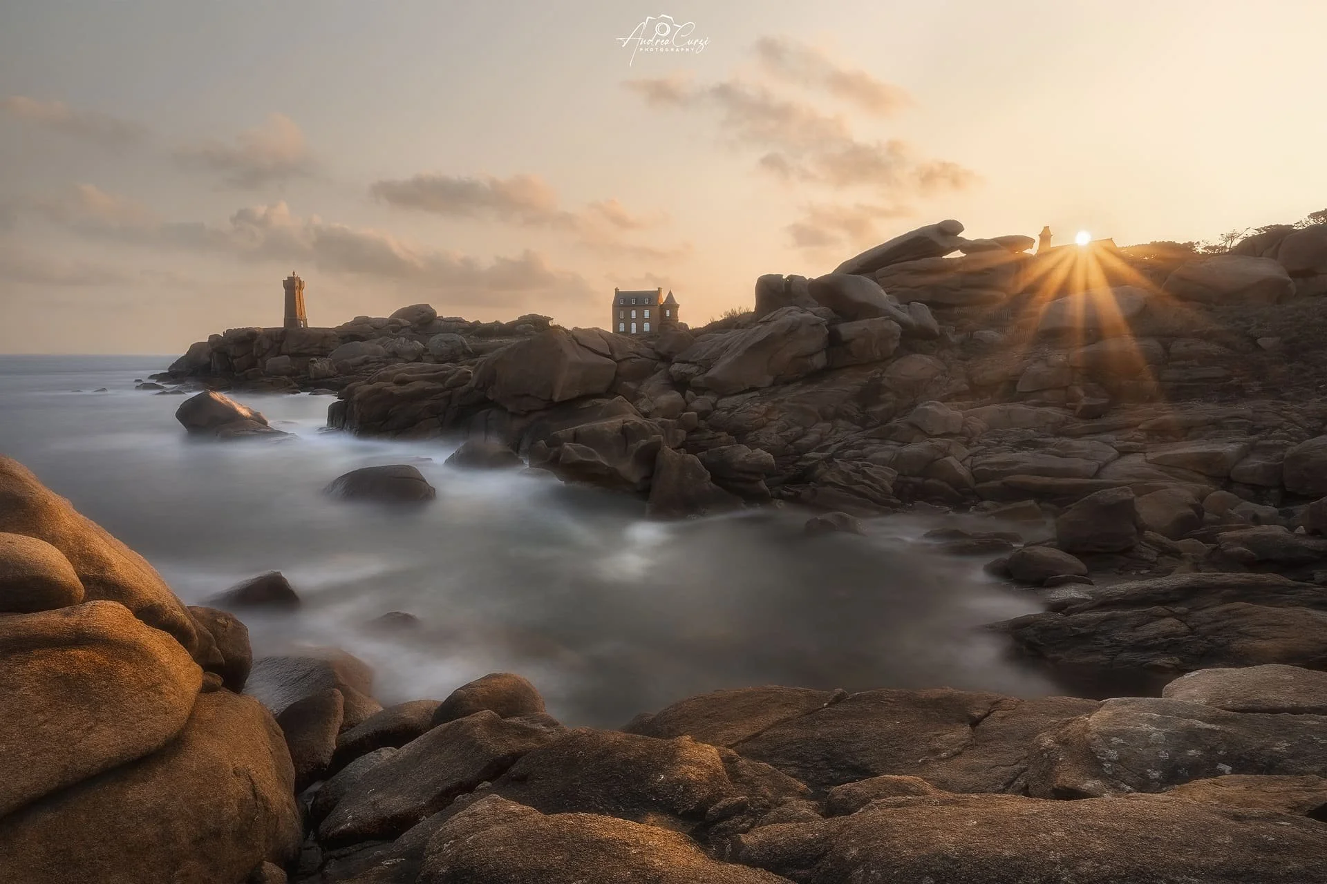 Faro di Ploumanac'h in Bretagna con costa di granito rosa all'alba, due torri, una di fronte all'altra, e il sole che albeggia all'orizzonte creando un effetto di raggi di luce tra le rocce.