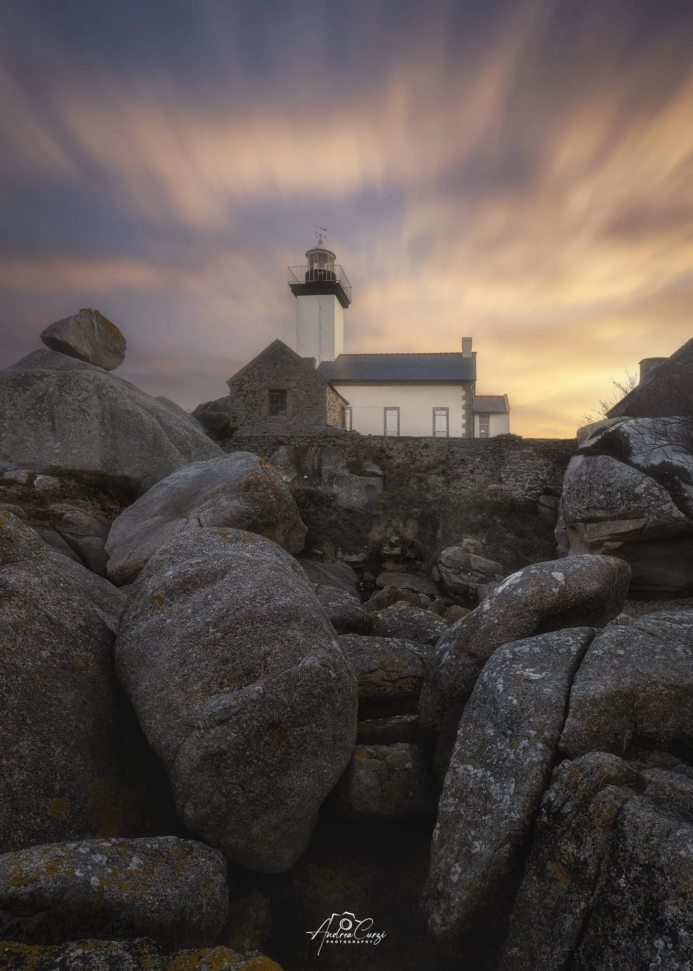 Faro di Pontusval in Bretagna su una scogliera con grandi rocce davanti, cielo al tramonto con nuvole allungate