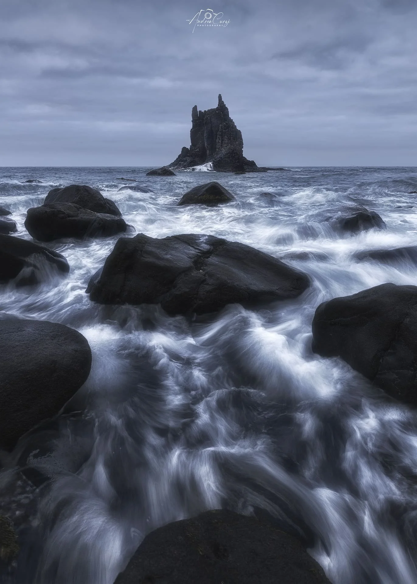Tramonto sulla spiaggia della Playa de benijo a Tenerife, con onde che si infrangono sui sassi e grandi scogli sullo sfondo, cielo scuro e nuvoloso.
