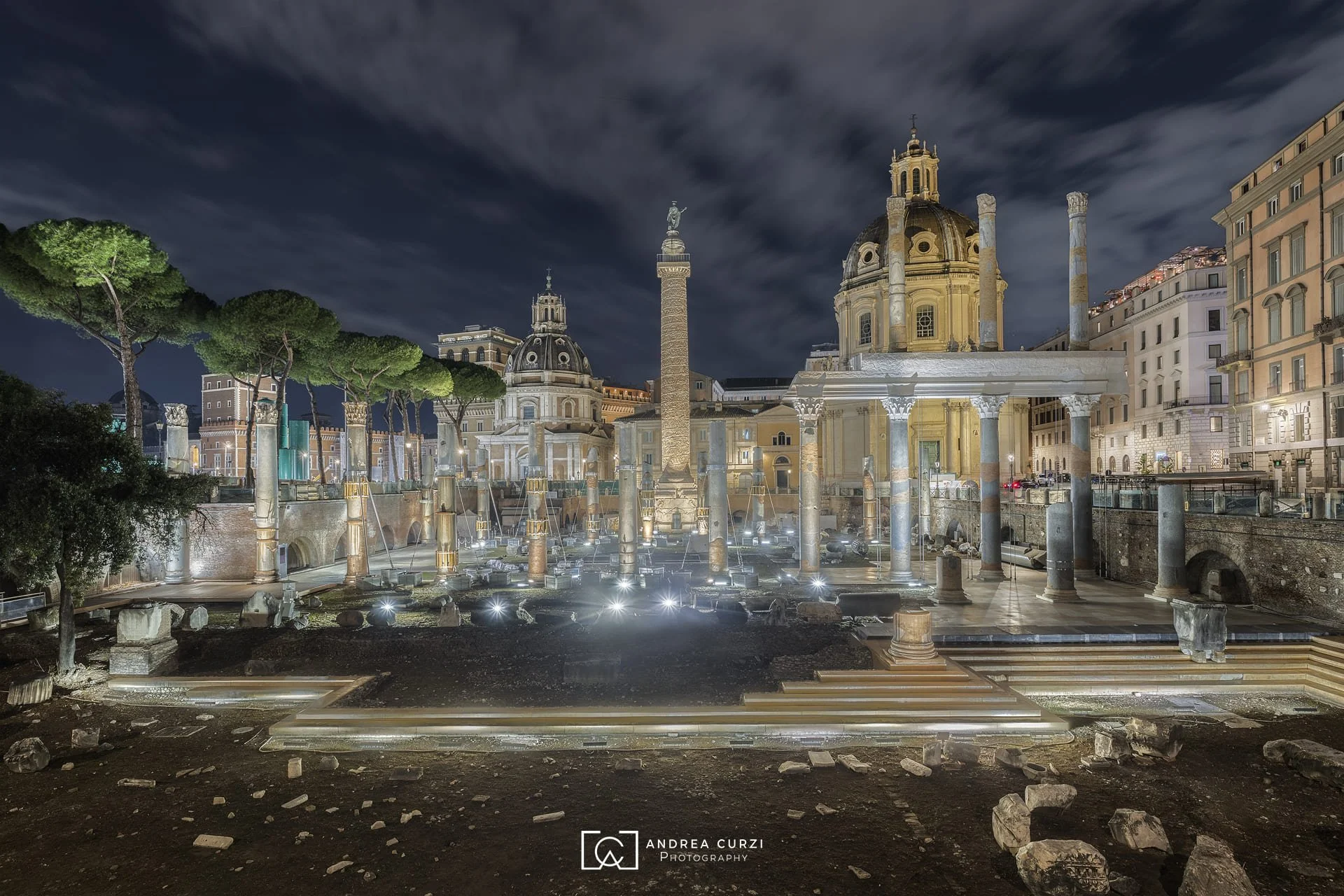 Vista notturna diei Fori di Traiano, con lampade che illuminano i resti archeologici e le chiese circostanti, sotto un cielo scuro.