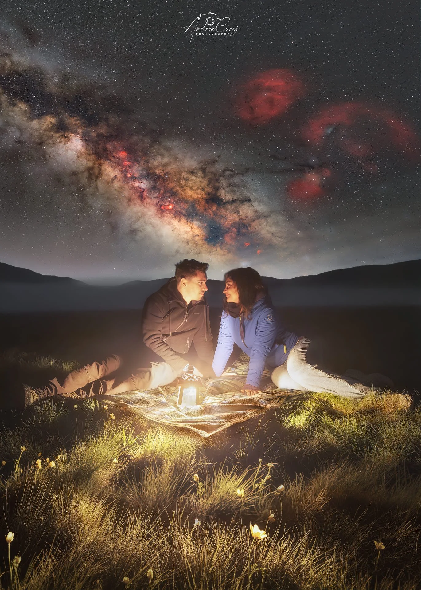 Due persone sedute su una coperta in un campo notturno a Castelluccio di Norcia in Umbria , illuminate da una lanterna, con il cielo stellato e la Via Lattea visibile sopra di loro.