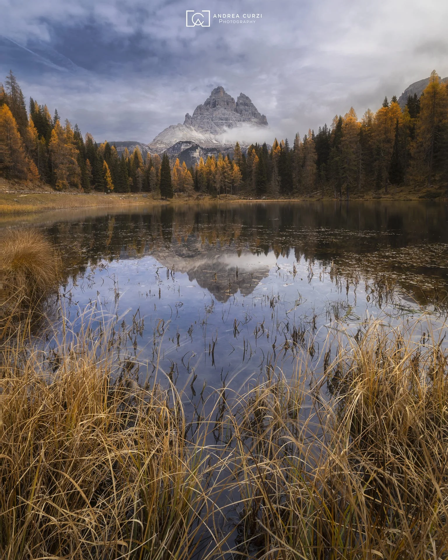 Paesaggio autunnale con un lago circondato da alberi colorati, montagne avvolte nella nebbia e cielo nuvoloso. La montagna più alta si riflette nell'acqua del lago.