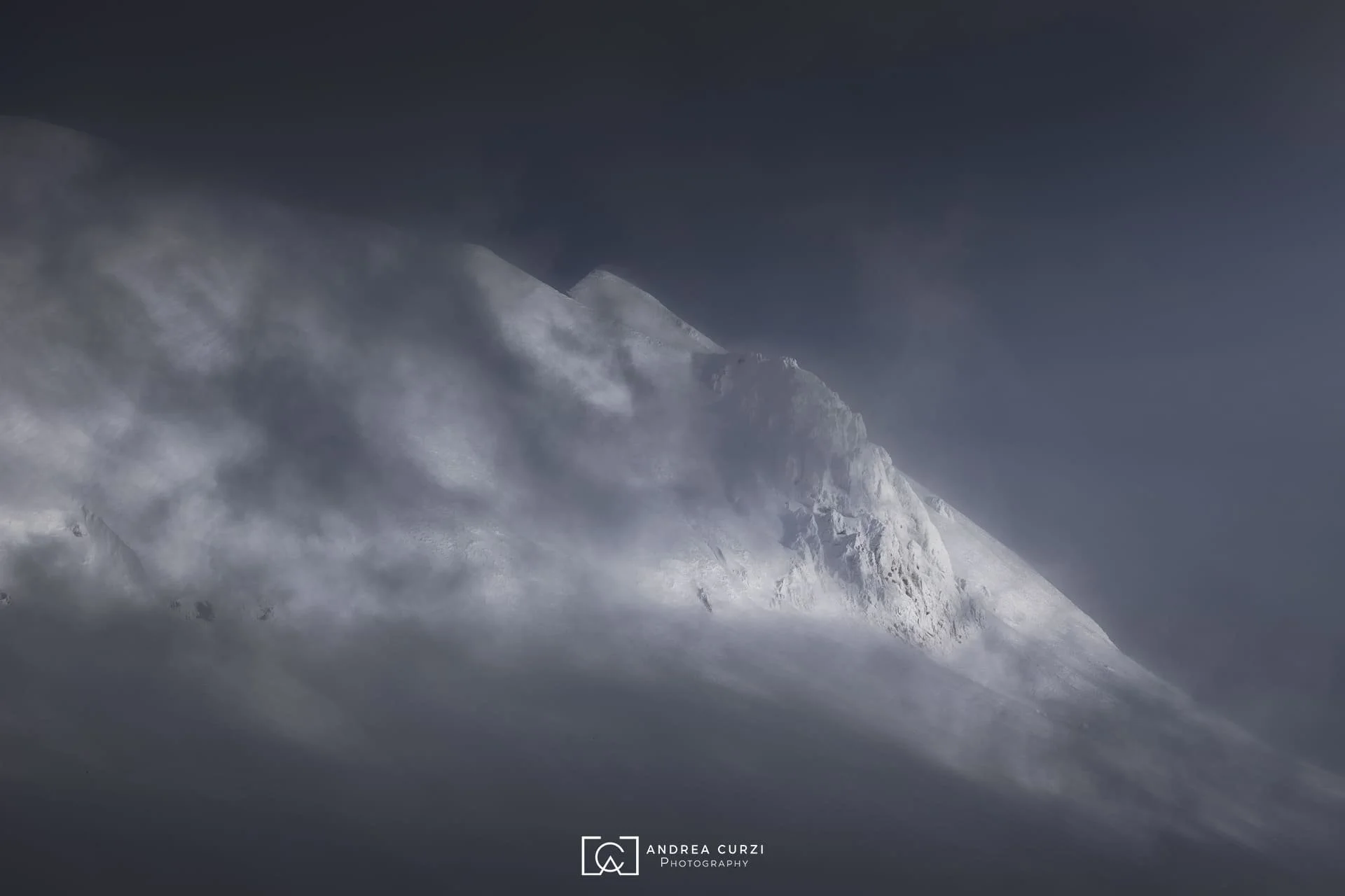Montagna innevata avvolta in nuvole e nebbia, con un cielo terso e scuro. Scattata sul Parco Nazionale dei Monti Sibillini a Castelluccio di Norcia durante un workshop fotografico di Andrea Curzi.