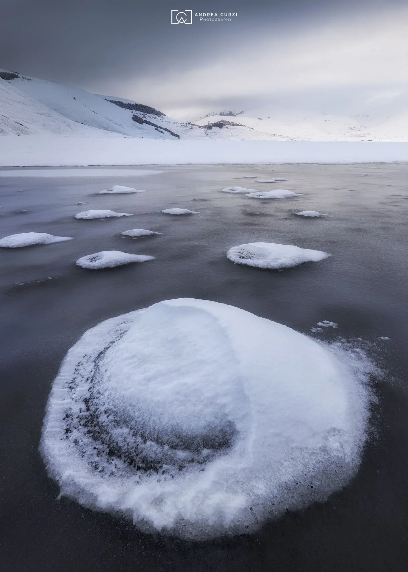 Paesaggio invernale con acqua ghiacciata e neve, montagne sullo sfondo e nuvole sopra. Scattata sul Parco Nazionale dei Monti Sibillini a Castelluccio di Norcia durante un workshop fotografico di Andrea Curzi.