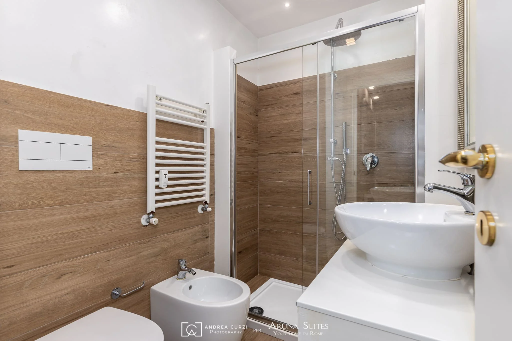 Bagno moderno di un'affittacamere con pareti in legno, doccia con porta in vetro, lavandino rotondo su mobile bianco, bidet e tenda riscaldamento bianca. Scattata durante un servizio fotografico di interni di Andrea Curzi nelle Marche