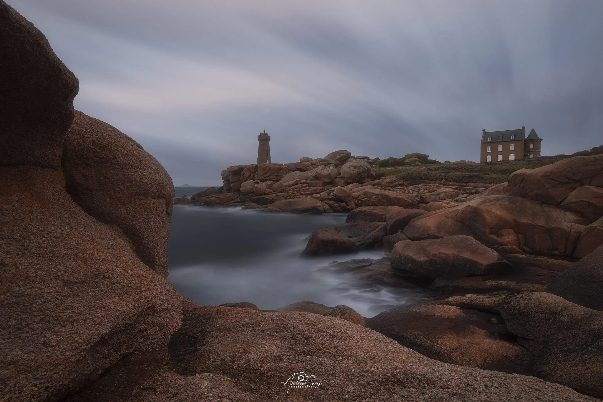 Faro di Ploumanac'h in Bretagna lungo la costa di granito rosa con onde che scorrono, torre e casa in lontananza sotto un cielo nuvoloso.