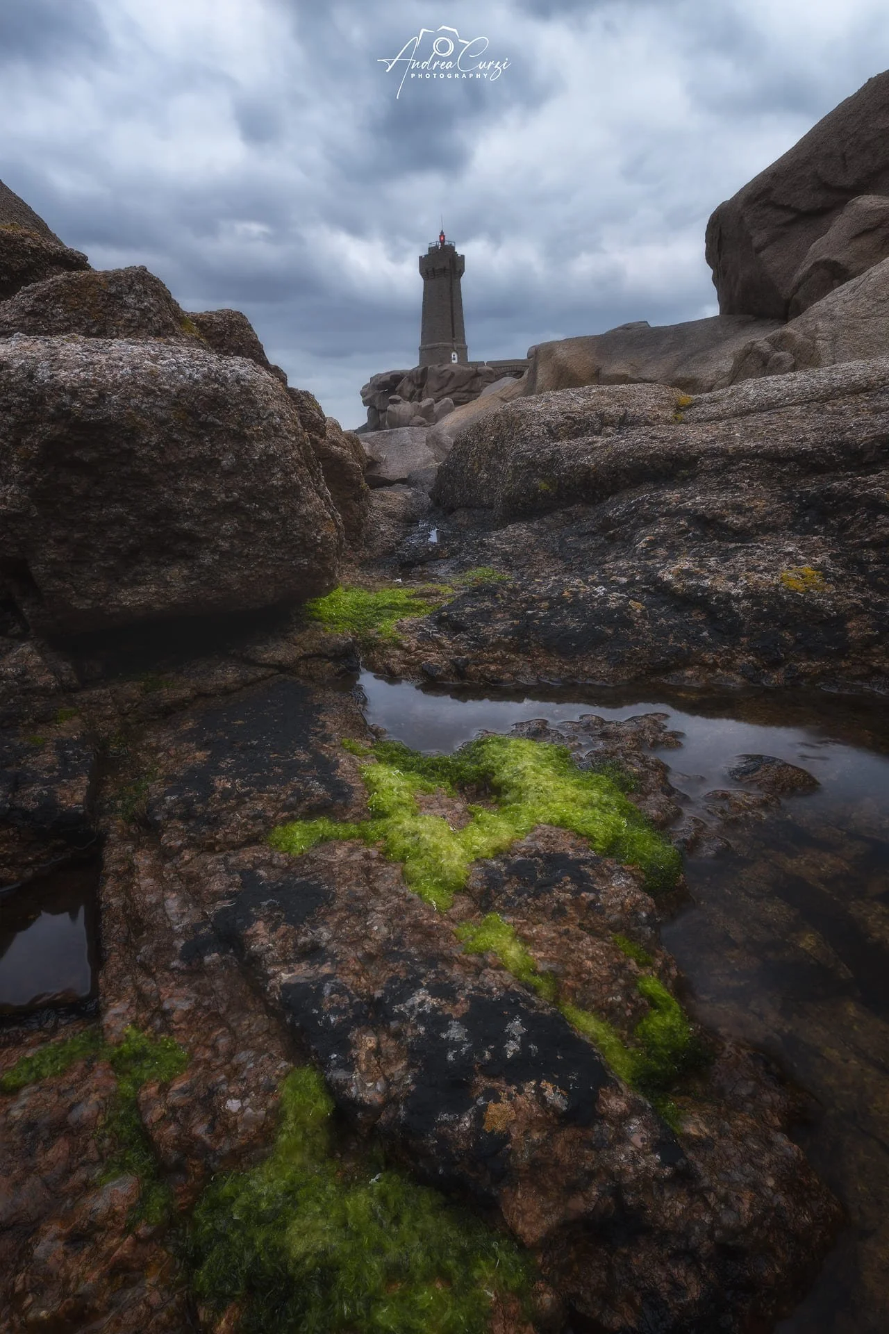 Faro di Ploumanac'h in Bretagna sulla cima di una scogliera, visto attraverso rocce ricoperte di muschio verde, sotto un cielo nuvoloso.