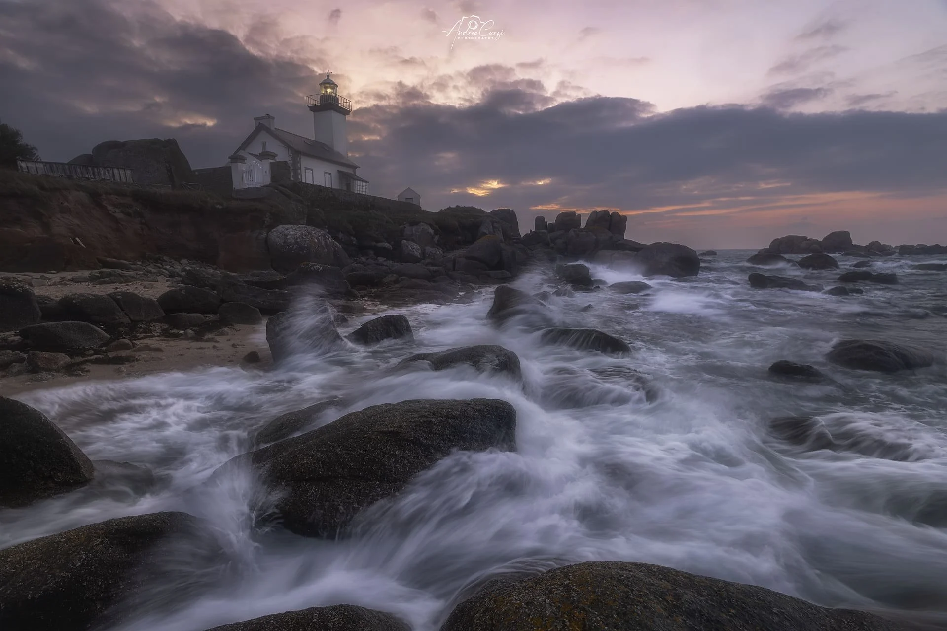 Scogliera sul faro di Pontusval in Bretagna con onde che si infrangono sulle rocce e un faro sulla collina al tramonto
