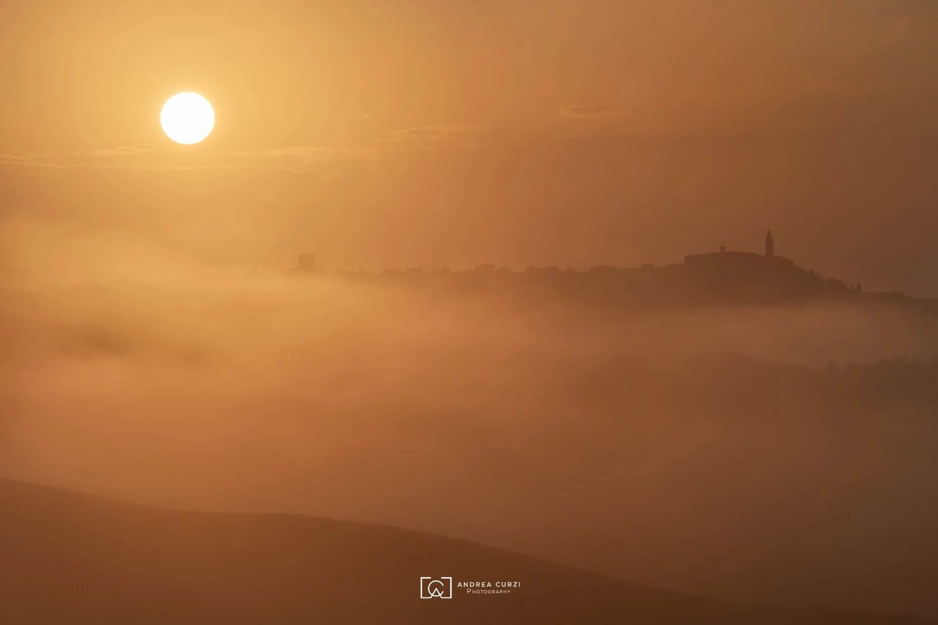 Sole all' alba che splende sopra una valle avvolta nella nebbia, con silhouette di colline e un campanile in lontananza. Scattata in Val d'Orcia in Toscana durante un workshop fotografico 1to1 di Andrea Curzi.