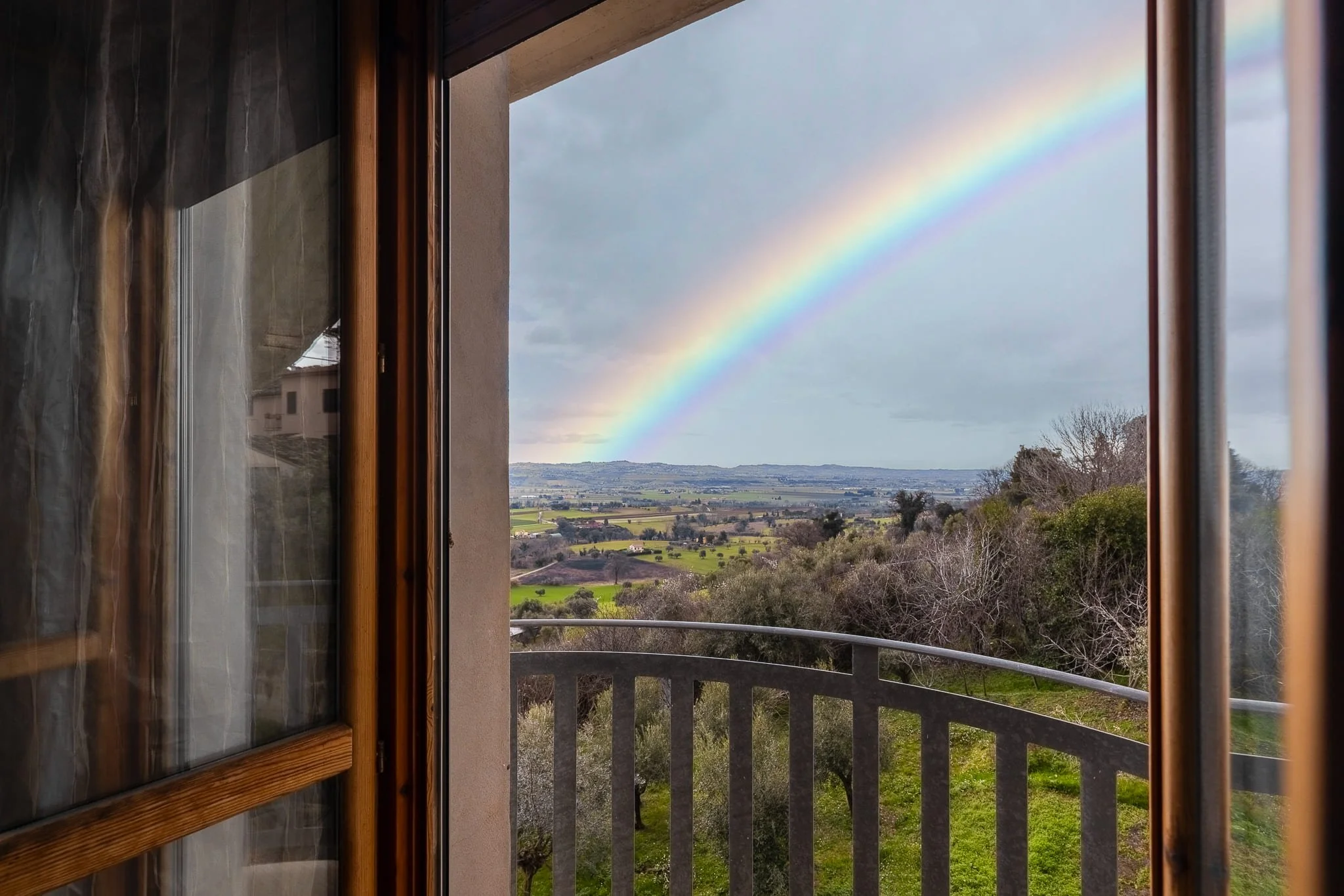 Vista di un arcobaleno doppio tra il cielo nuvoloso e un paesaggio rurale con campi verdi e alberi, vista da una finestra di un B&B. Scattata durante un servizio fotografico di interni di Andrea Curzi nelle Marche