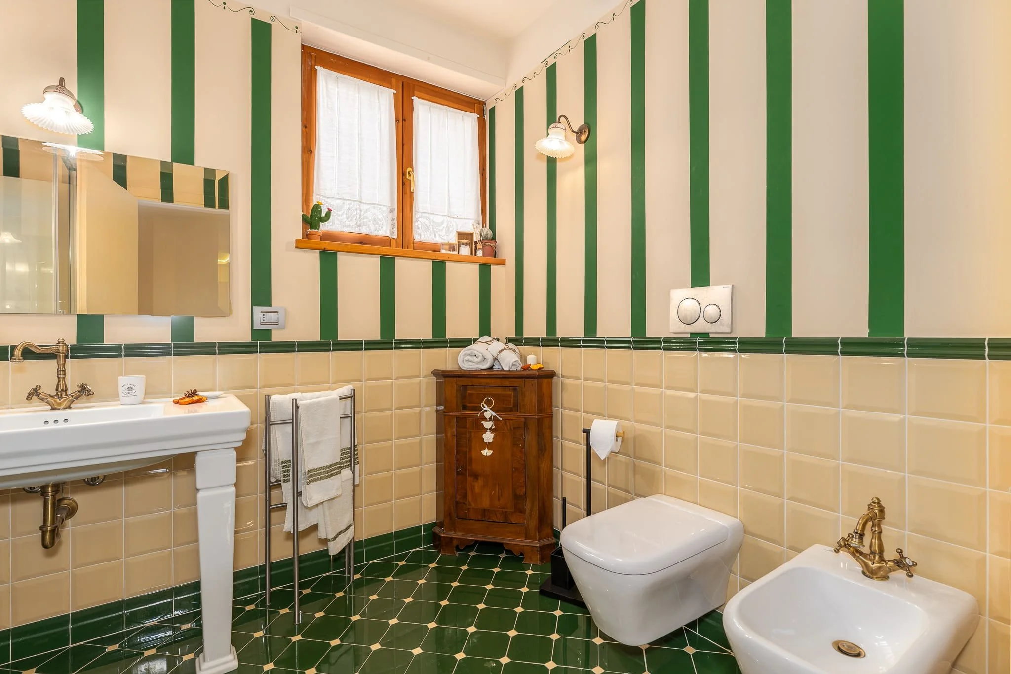 Bagno con pareti a righe verdi e crema di un B&B, lavabo, specchio, finestra con tende, armadietto in legno, toilette e bidet con accessori in ottone. Scattata durante un servizio fotografico di interni di Andrea Curzi nelle Marche