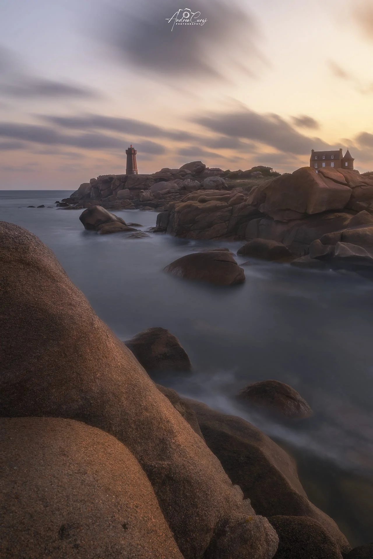 Faro di Ploumanac'h in Bretagna con grandi massi rossi e un faro sulla riva, all'alba, con nuvole veloci nel cielo e la costa di granito rosa.