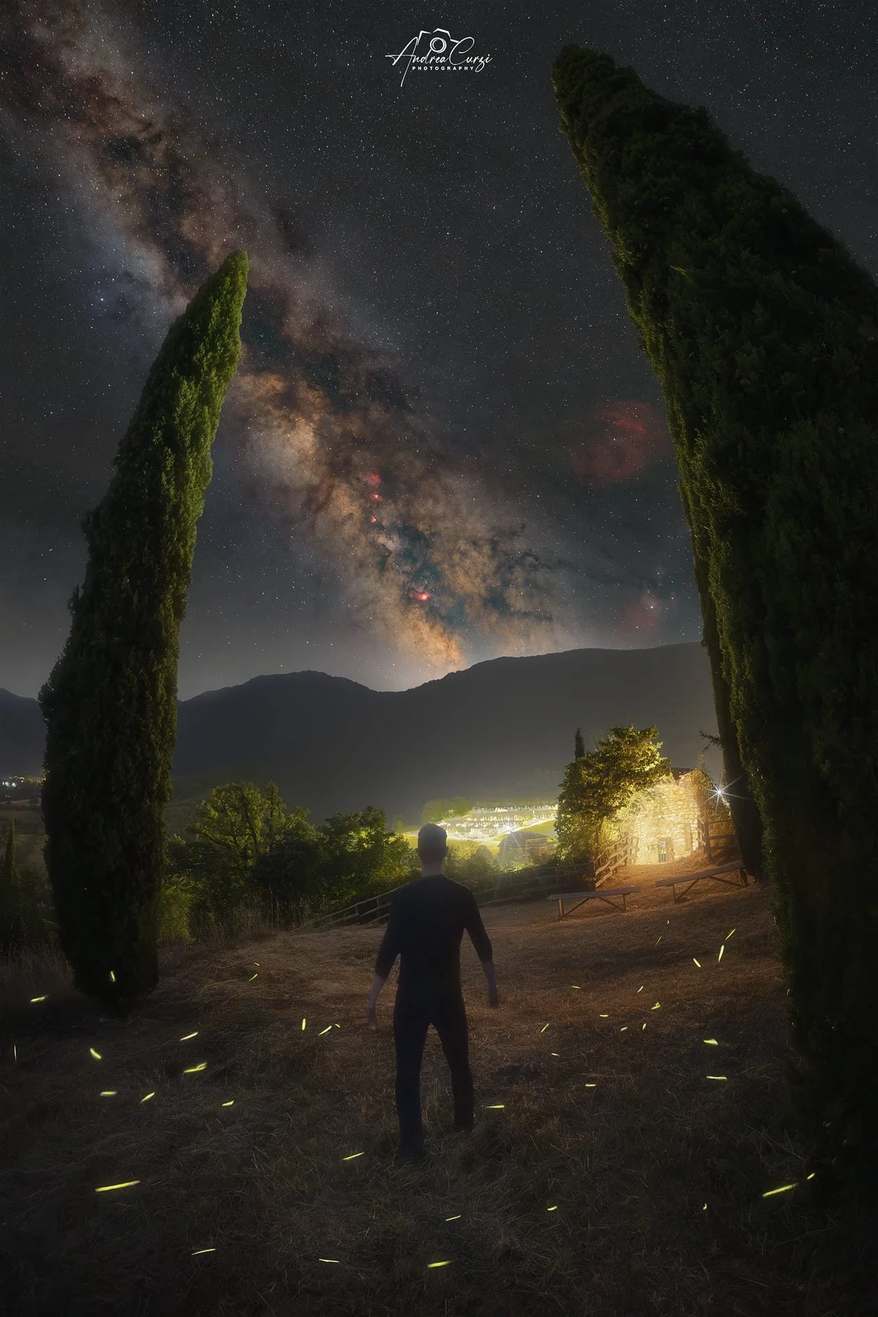 Una persona silhouette si trova in un campo con due grandi cipressi a sinistra e a destra, sotto un cielo notturno con la Via Lattea visibile, montagne sullo sfondo, e una piccola casa illuminata a destra. Siamo sui Monti Sibillini, sul lago di Fiast