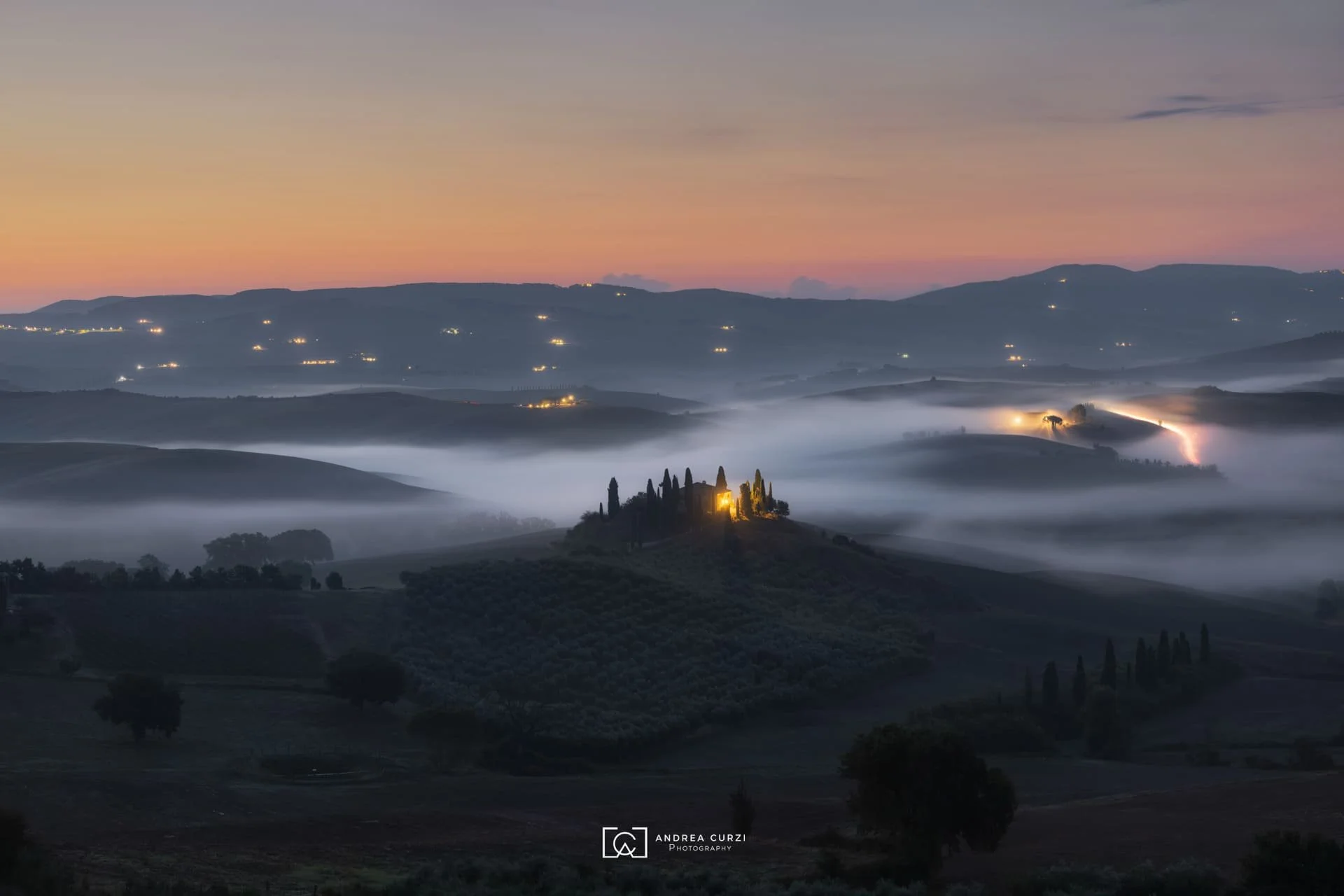 Paesaggio collinare del Podere Belvedere all'alba con nebbia e luci sparse, alberi di cipresso e una casa illuminata su una collina. Scattata in Val d'Orcia in Toscana durante un workshop fotografico 1to1 di Andrea Curzi.
