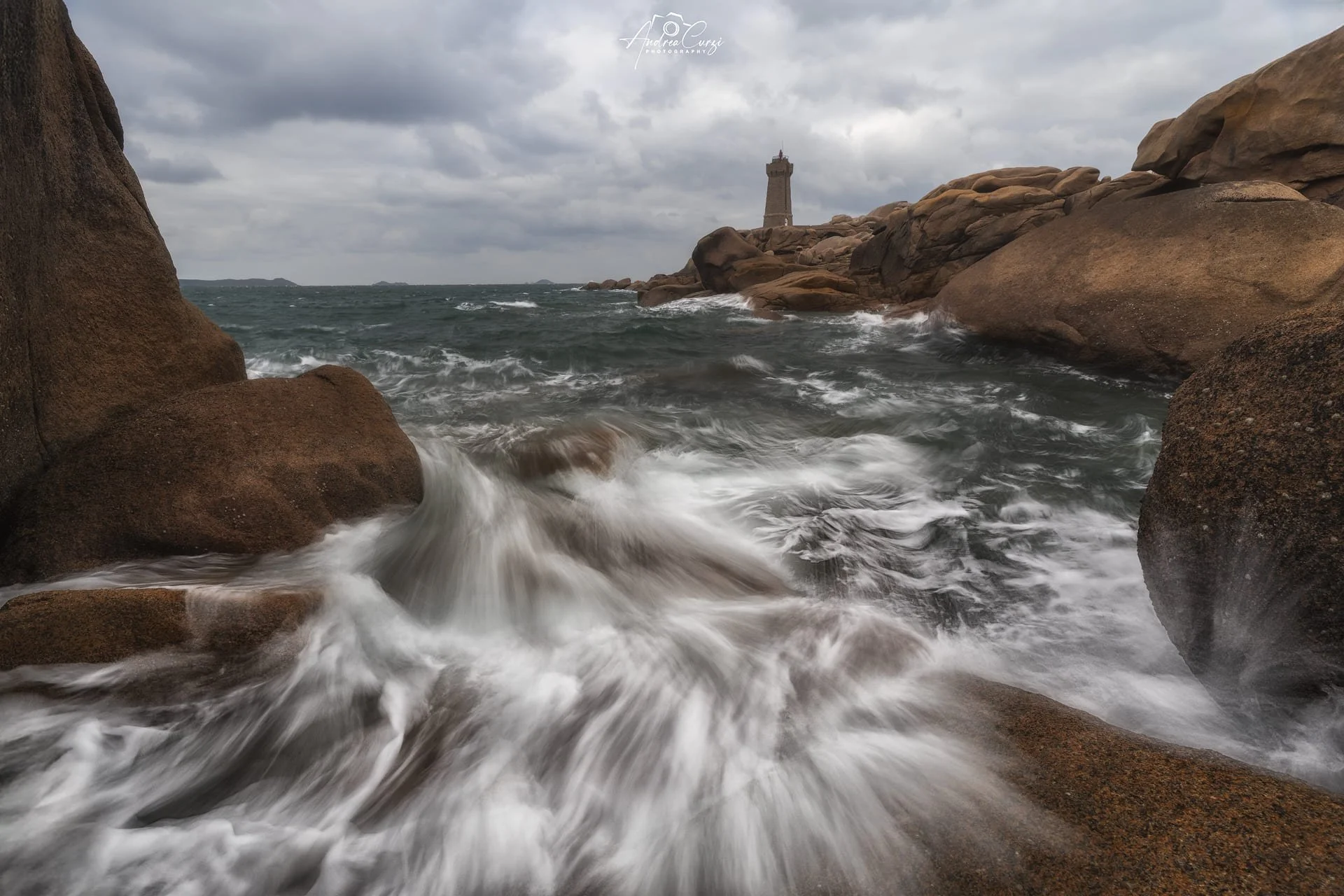 Faro di Ploumanac'h in Bretagna con onde che si infrangono sulla costa di granito rosa e un faro in lontananza sotto un cielo nuvoloso.