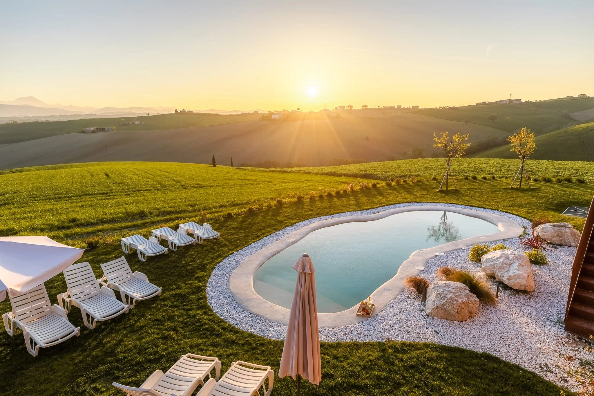 Piscina rotonda di un casolare con sdraio bianche, ombrelloni e paesaggi collinari al tramonto. Scattata durante un servizio fotografico di interni di Andrea Curzi nelle Marche