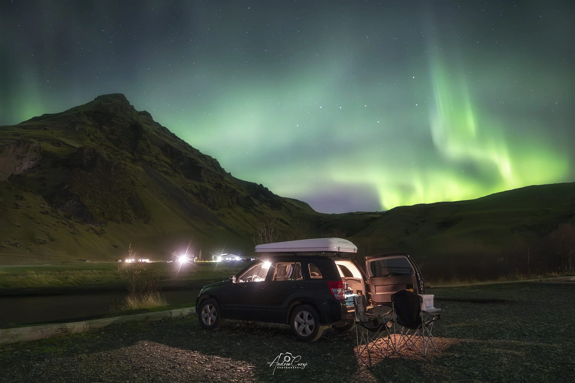 Auto nera con tenda da tetto e sedie pieghevoli sul primo piano, sotto il cielo stellato con le aurore boreali verdi e viola sopra montagne verdi in Islanda.