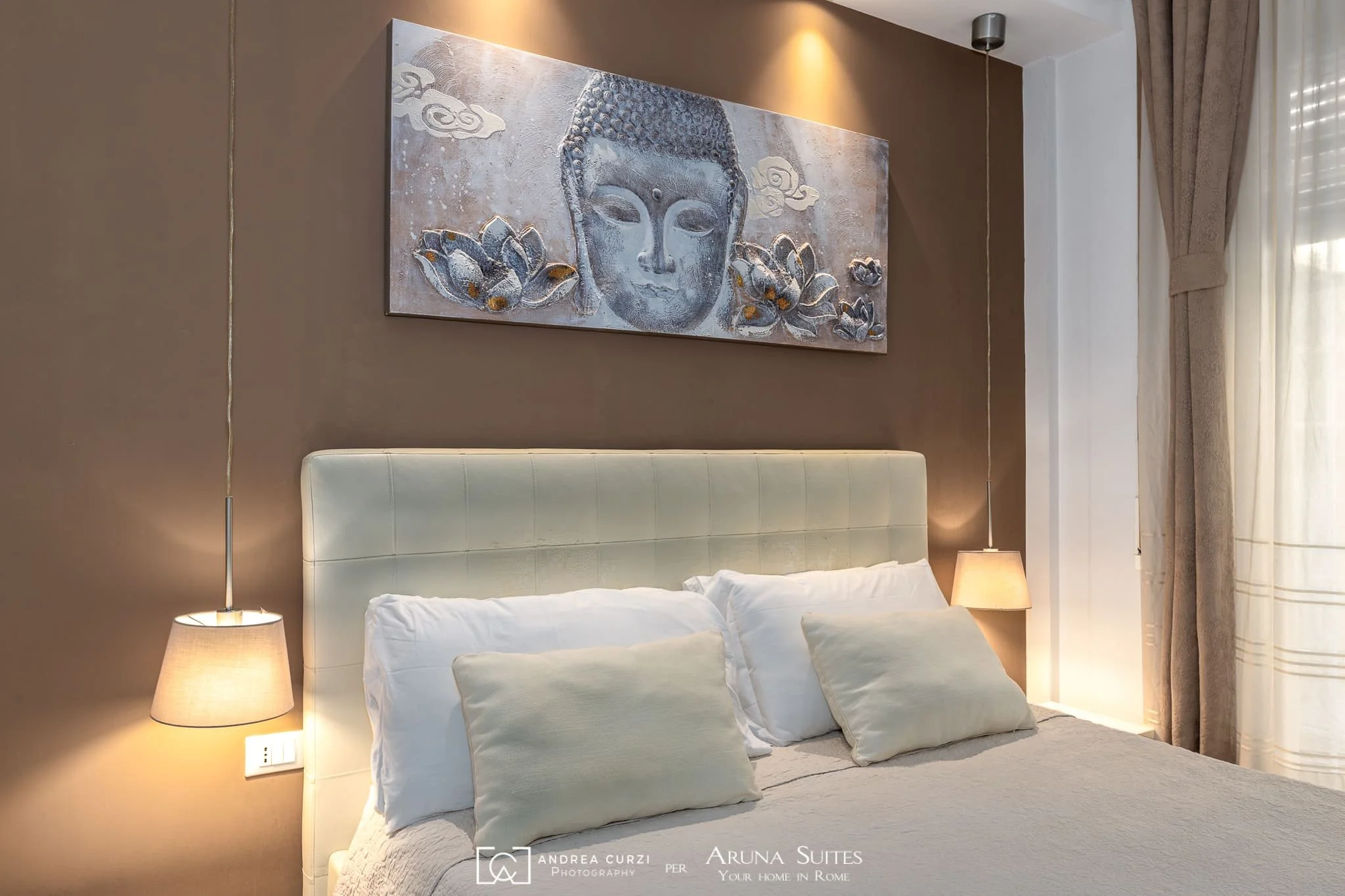 Letto matrimoniale di un'affittacamere con abat-jour ai lati, quadro con volto di Buddha e fiori sulla parete sopra il letto, tende chiare in fondo alla stanza. Scattata durante un servizio fotografico di interni di Andrea Curzi nelle Marche