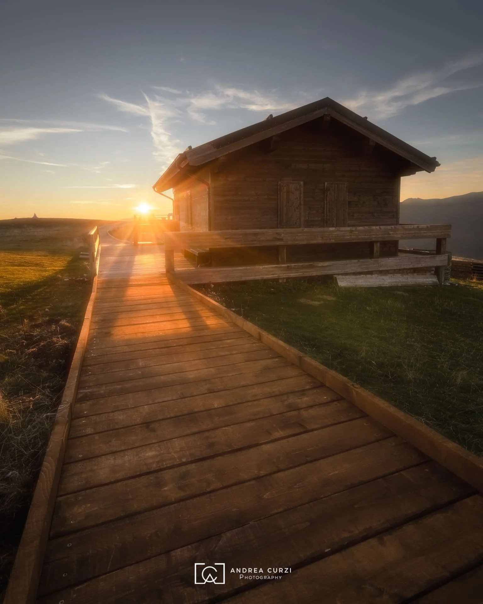 Casa di legno con passerella in legno che conduce verso il tramonto in un paesaggio rurale montano. Scattata sul Parco Nazionale dei Monti Sibillini nelle Marche durante un workshop fotografico di Andrea Curzi.