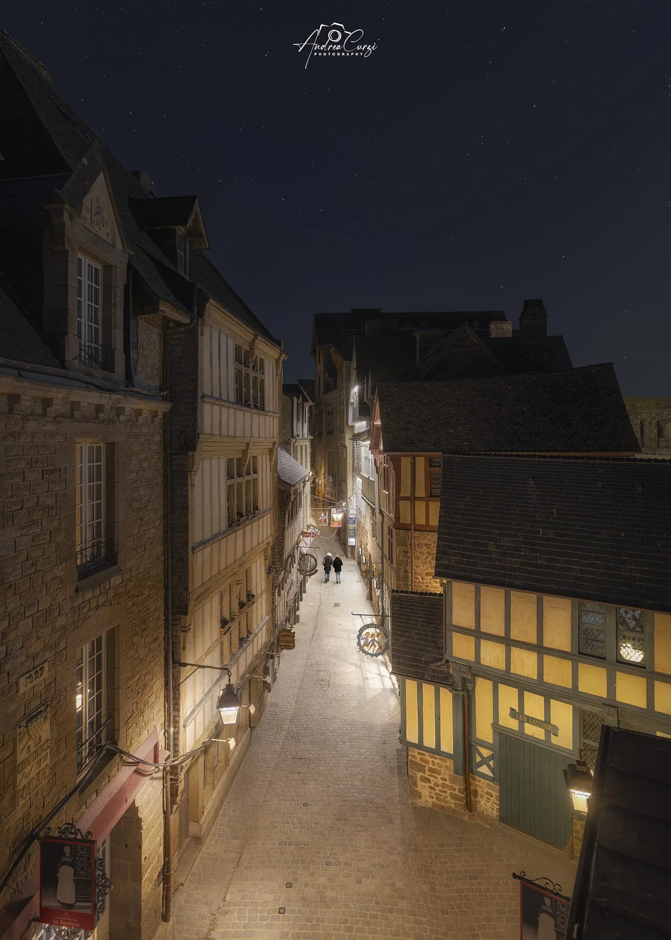 Castello di Mont Saint Michel in Normandia di notte con un cielo stellato, visibile dal basso tra edifici medievali in pietra e due persone che passeggiano