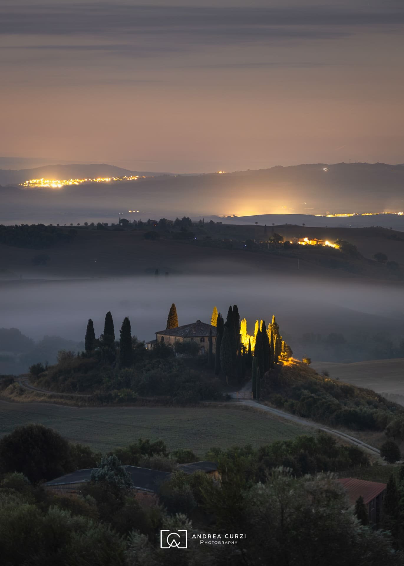 Paesaggio notturno del Podere Belvedere con colline, alberi e una casa illuminata al centro, circondata da nebbia, con città illuminate sullo sfondo. Scattata in Val d'Orcia in Toscana durante un workshop fotografico 1to1 di Andrea Curzi.