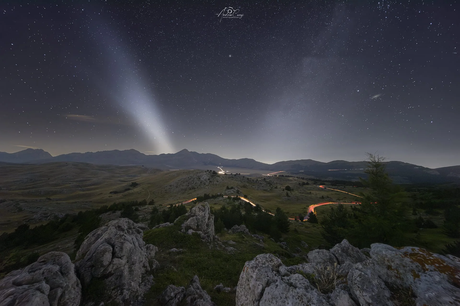 Paesaggio notturno con montagne, rocce, alberi, scie delle auto, il cielo stellato, la coda della via lattea e la scia di gas del razzo Falcon 9. Siamo a Campo imperatore in Abruzzo.