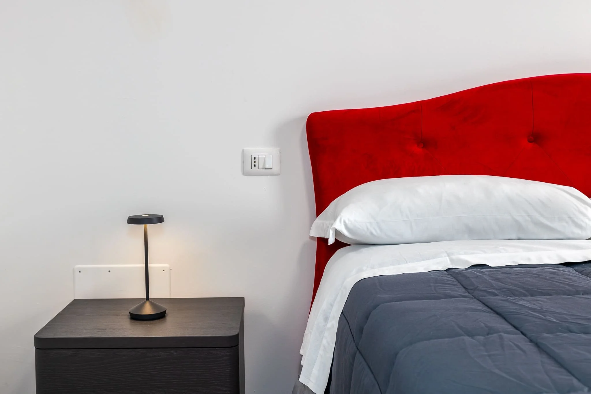 Letto matrimoniale di un'affittacamere, cuscino bianco e biancheria grigia, accanto a un comodino nero con lampada nera, parete bianca con presa elettrica. Scattata durante un servizio fotografico di interni di Andrea Curzi nelle Marche