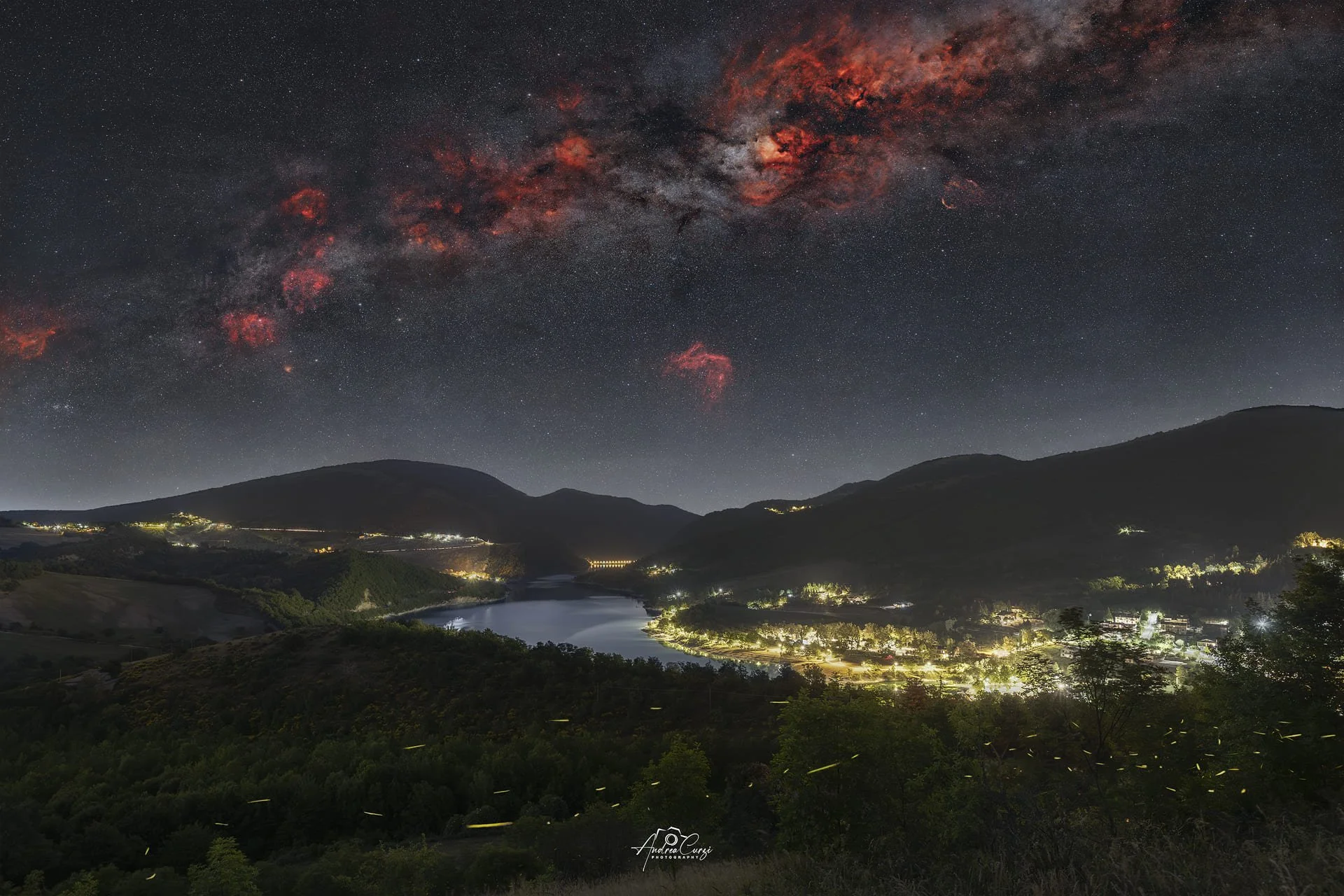 Paesaggio notturno con montagne, il Lago di Fiastra e il paese illuminato dal basso, sotto un cielo stellato e la coda della Via Lattea con nebulose rosse e galassie. Siamo sui Monti Sibillini, sul lago di Fiastra.