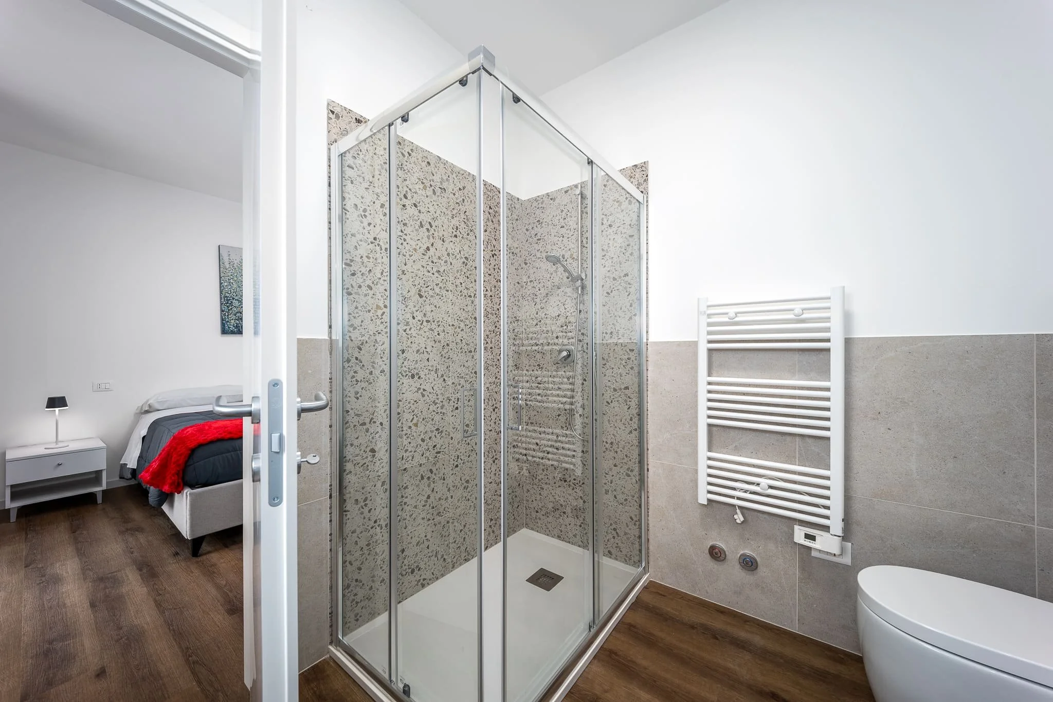 Bagno di un'affittacamere con cabina doccia, termo riscaldato bianco, pareti beige e pavimento in legno. Scattata durante un servizio fotografico di interni di Andrea Curzi nelle Marche