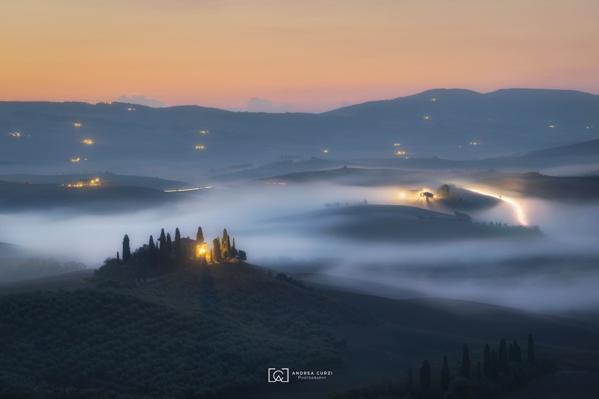 Paesaggio collinare del Podere Belvedere all'alba con nuvole di nebbia, alcune case illuminate e strade illuminate tra le colline. Scattata in Val d'Orcia in Toscana durante un workshop fotografico 1to1 di Andrea Curzi.