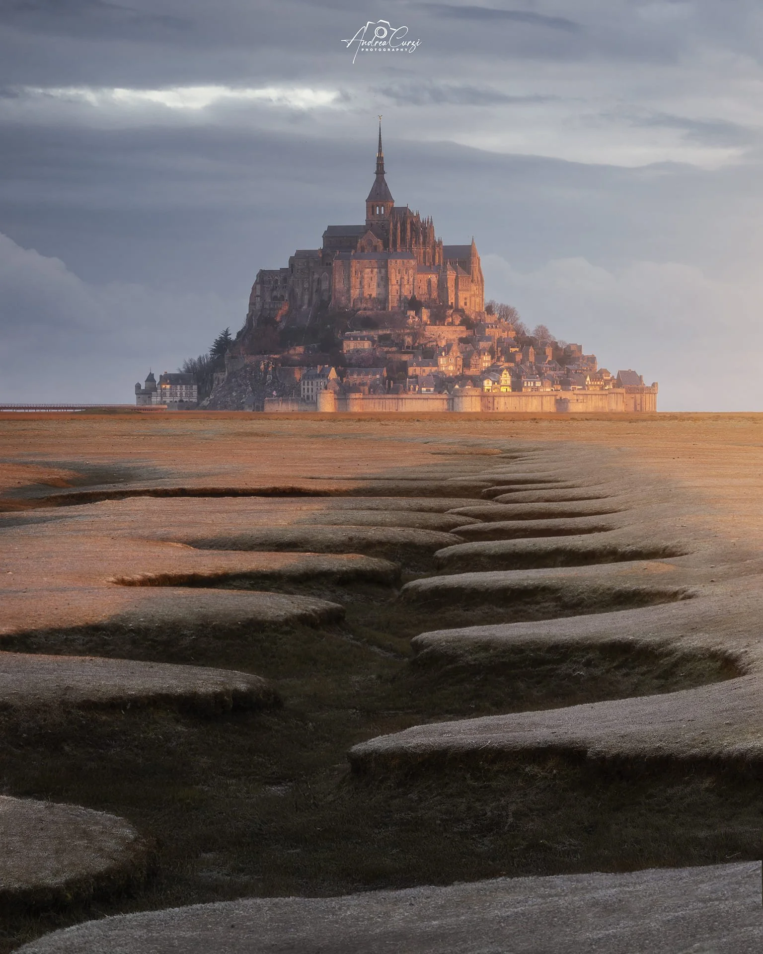Mont Saint-Michel con le sue antiche mura e la chiesa che si erge sopra un'isola rocciosa, visibile da lontano, con il classico serpentone in primo piano all'alba con cielo nuvoloso.