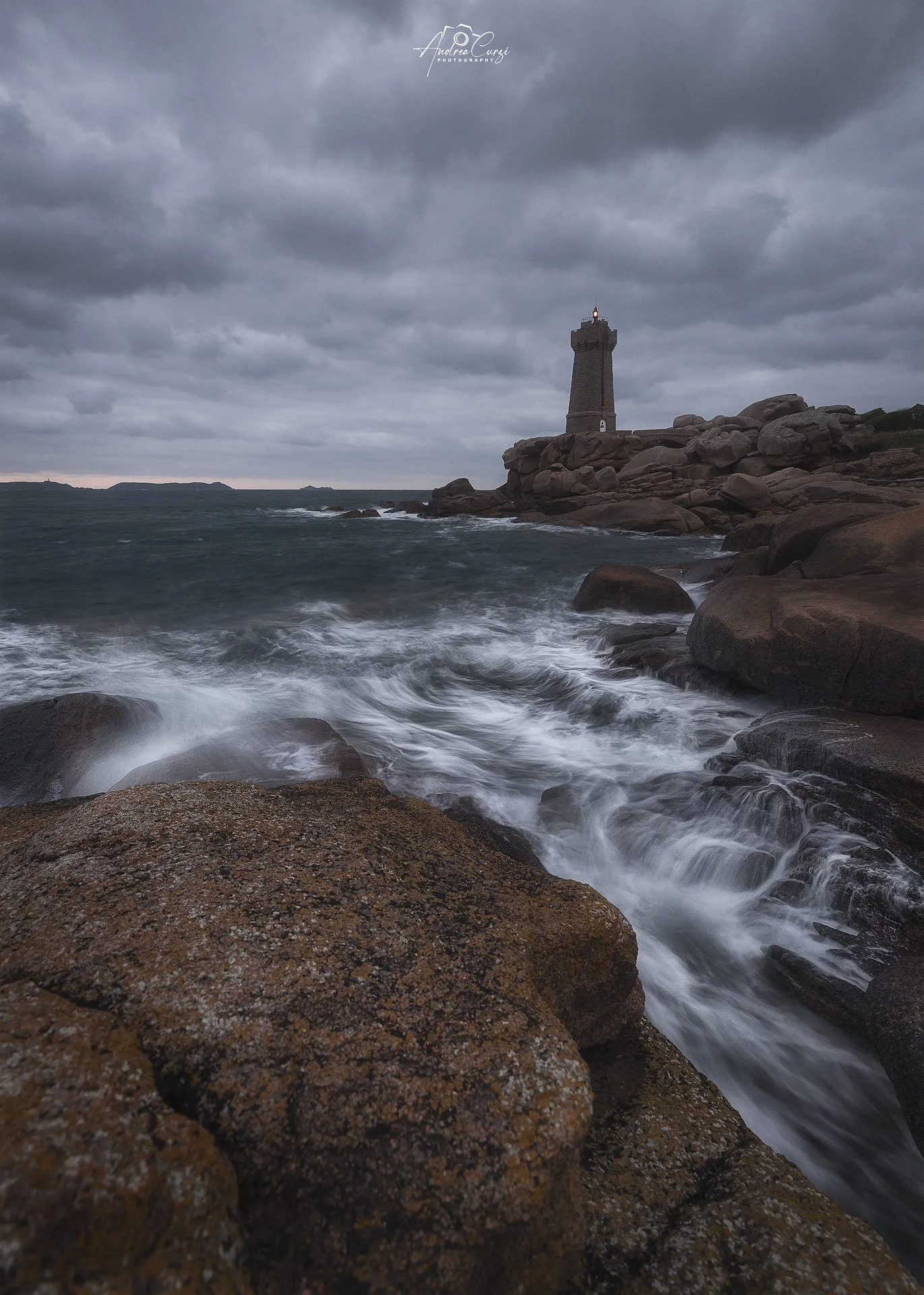 Faro di Ploumanac'h in Bretagna sulla costa con un faro in lontananza sotto un cielo nuvoloso, onde che si infrangono contro le rocce.