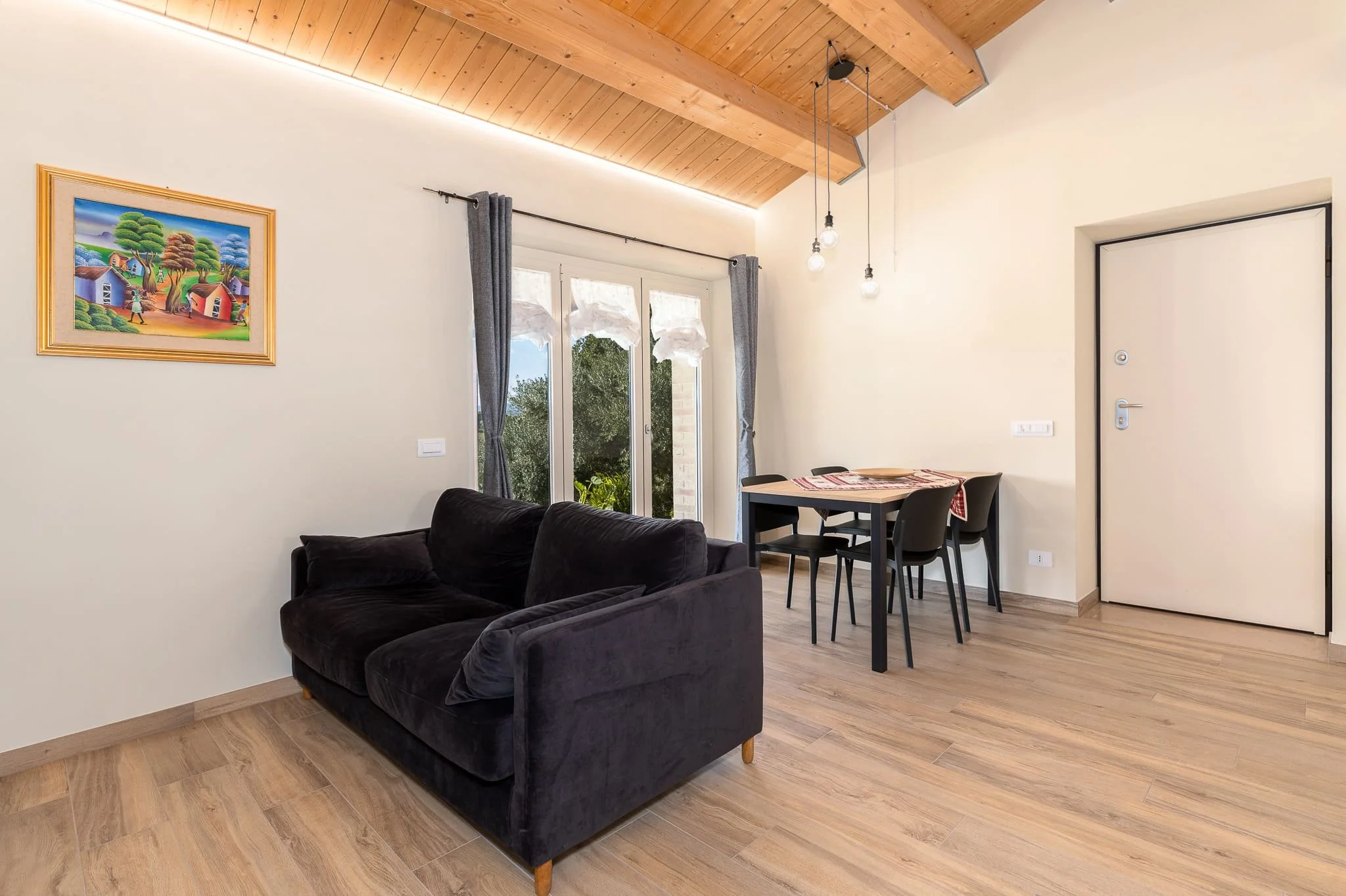Interno di un soggiorno di un B&B con divano nero, tavolo da pranzo con sedie nere, pareti bianche, pavimento in legno chiaro, finestra con tende grigie. Scattata durante un servizio fotografico di interni di Andrea Curzi nelle Marche