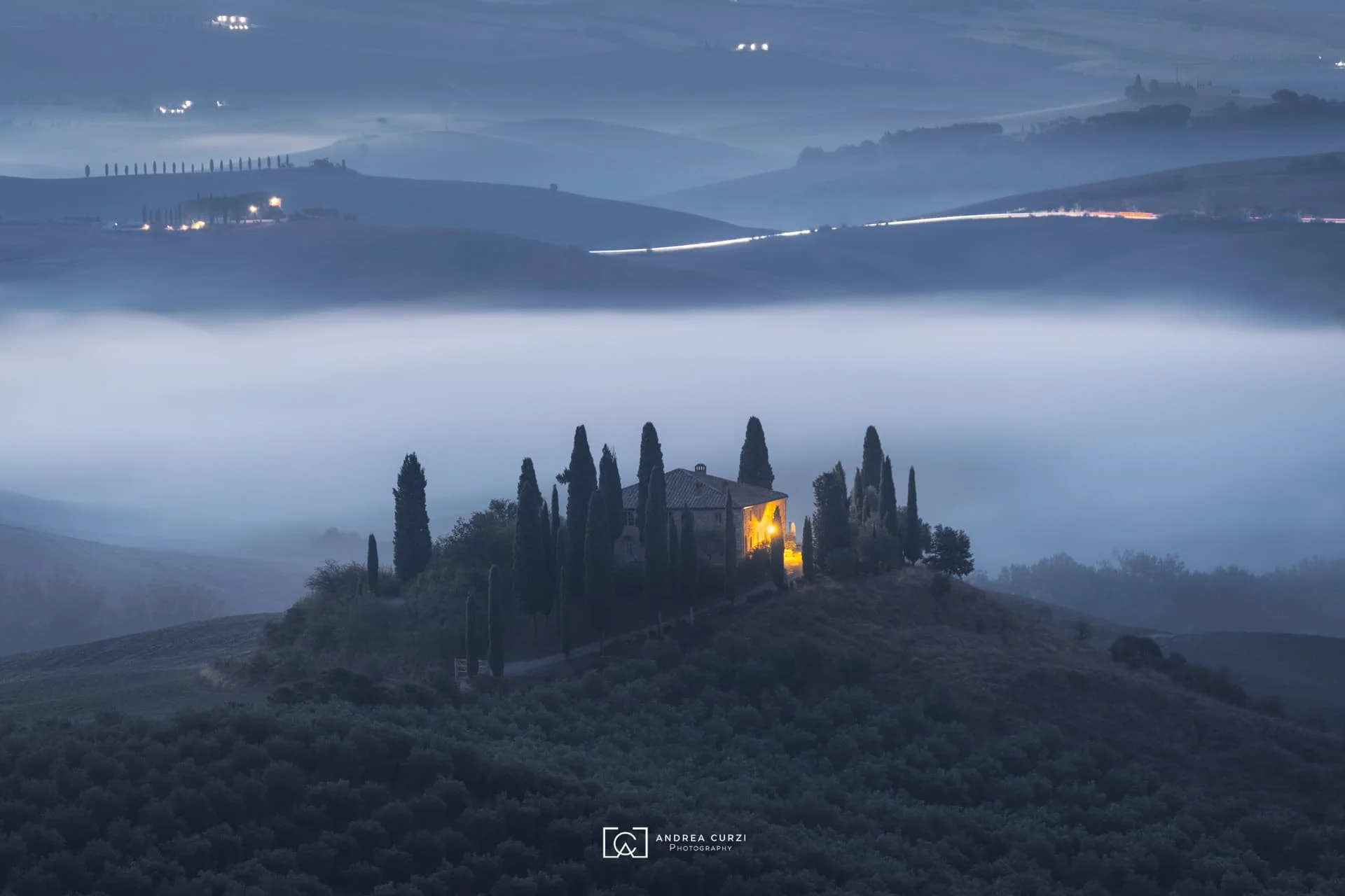 Paesaggio sereno del Podere Belveder con una casa illuminata circondata da cipressi su una collina, con nebbia e colline in lontananza, durante la sera o l'alba. Scattata in Val d'Orcia in Toscana durante un workshop fotografico 1to1 di Andrea Curzi.