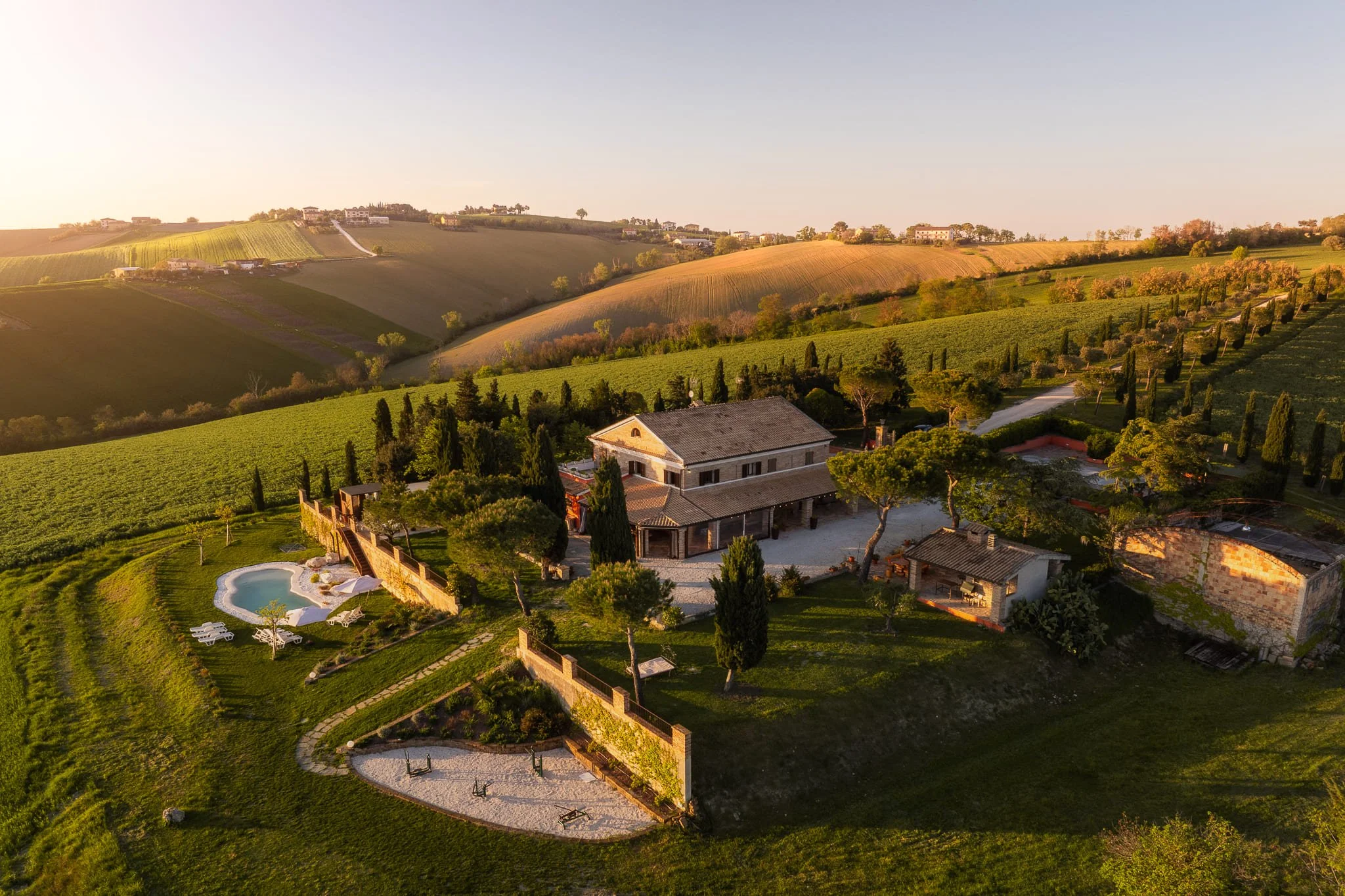 Paesaggio rurale con un casolare in pietra, piscina, alberi e campi verdissimi sul calar del sole. Scattata durante un servizio fotografico di interni di Andrea Curzi nelle Marche