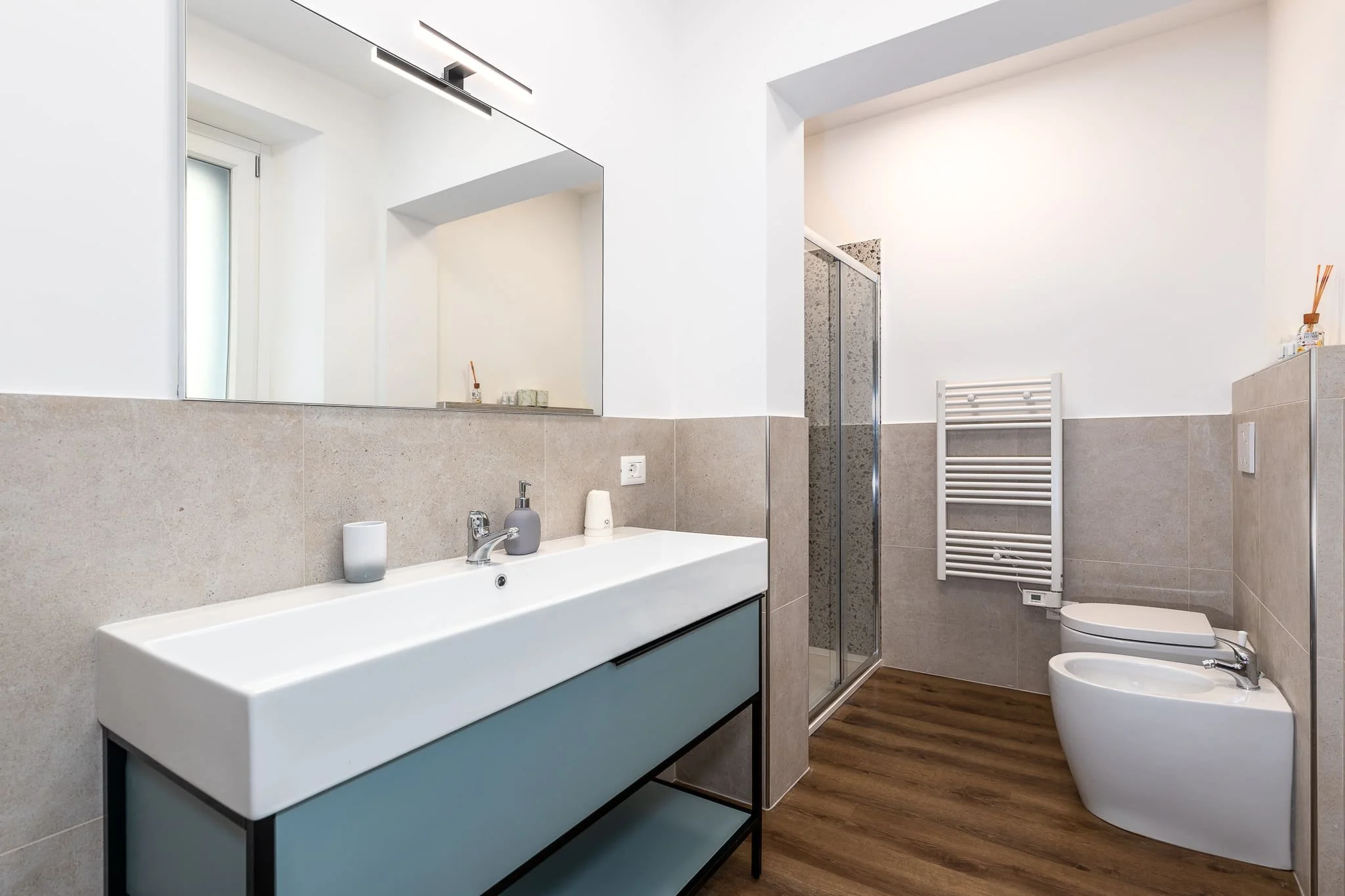 Bagno moderno di un affittacamere con lavandino, specchio grande, doccia con porta in vetro, water e bidet, asciugamano a riscaldamento e finestra. Scattata durante un servizio fotografico di interni di Andrea Curzi nelle Marche