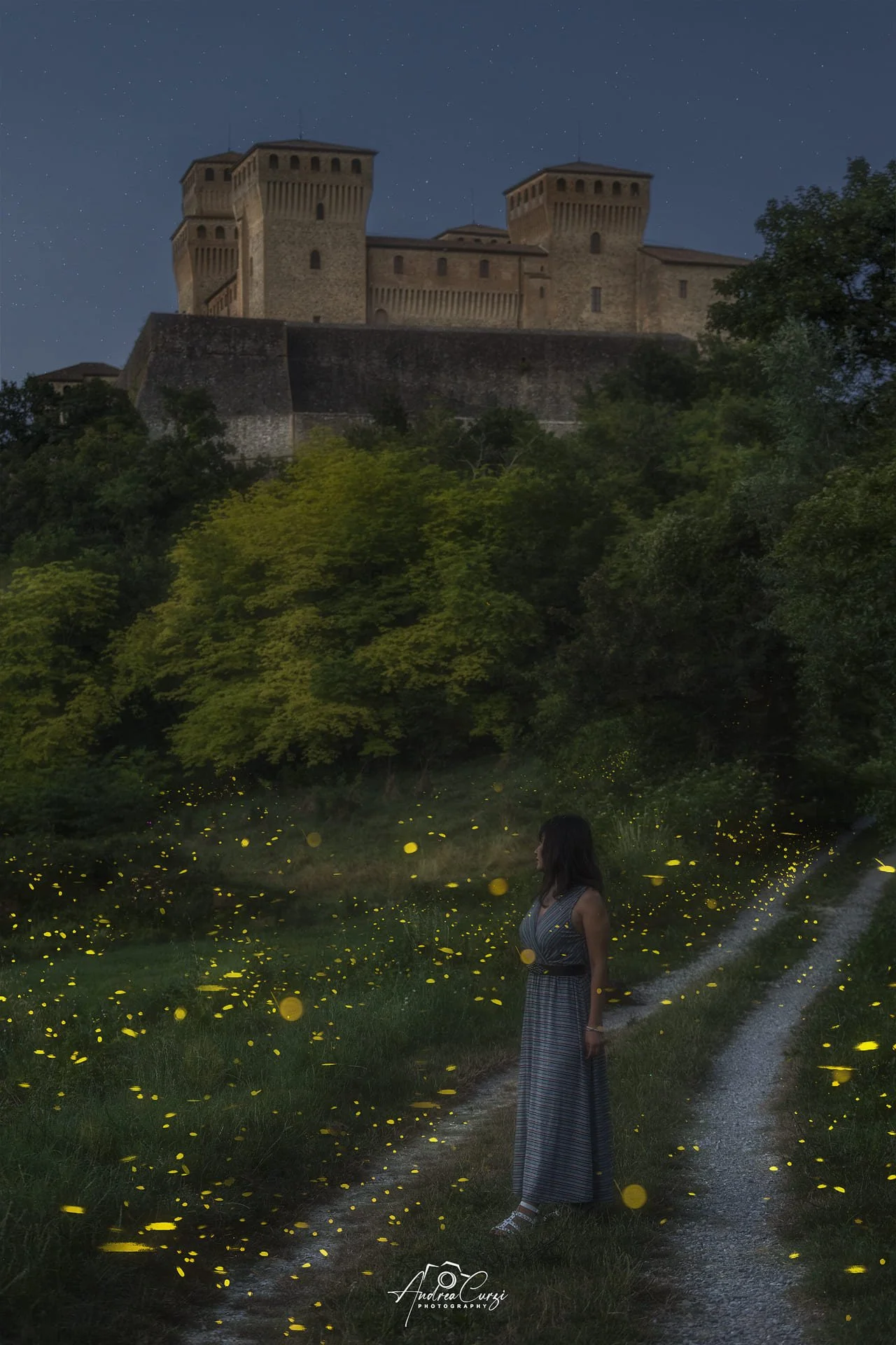 Una donna in abito lungo un sentiero in un bosco durante la notte, con il castello di Torrechiara sullo sfondo e un tappeto di lucciole luminose.