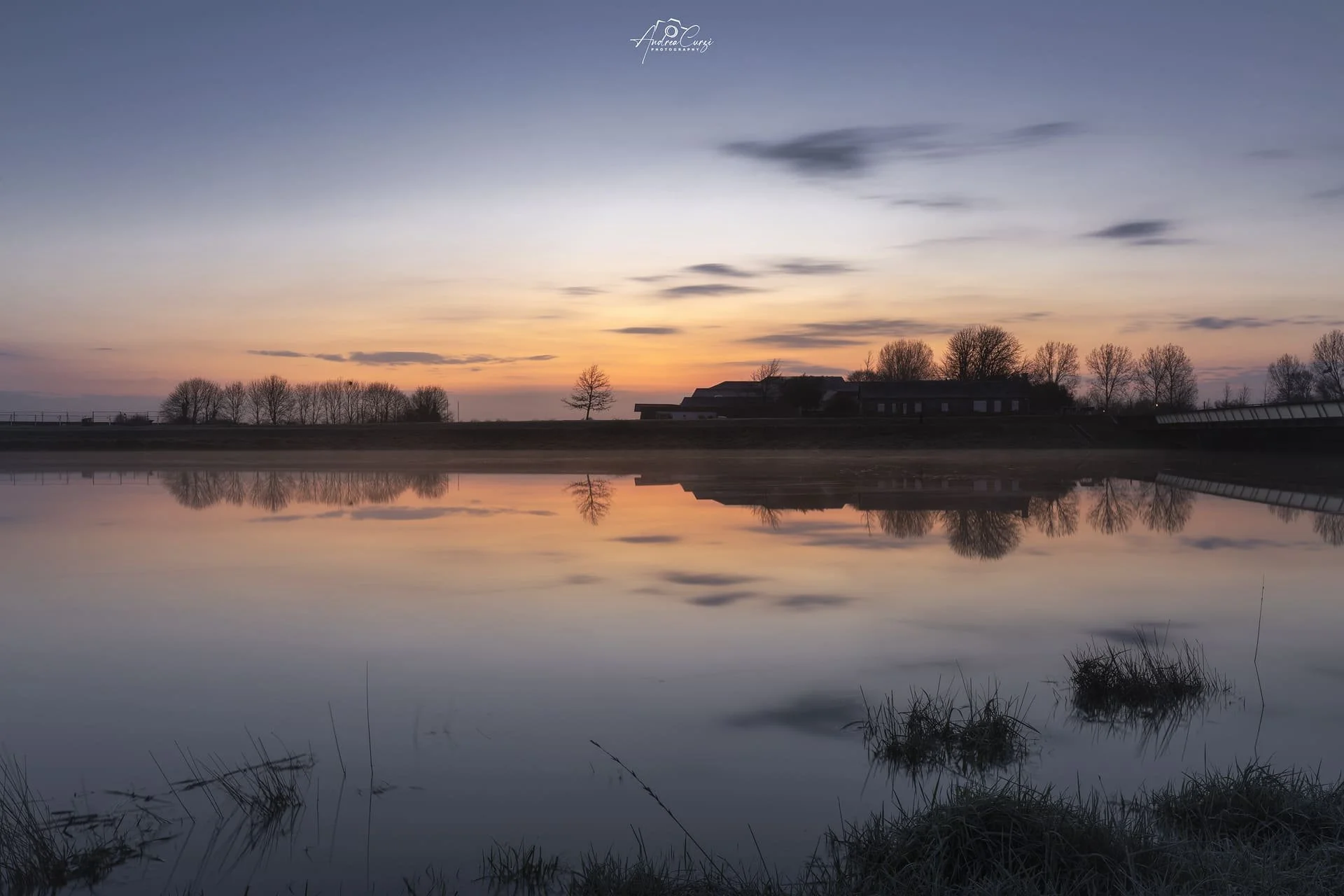 Tramonto con cielo blu e nuvole leggere, silhouette di alberi e edifici sullo sfondo, e riflesso dell'immagine nell'acqua calma di un lago.