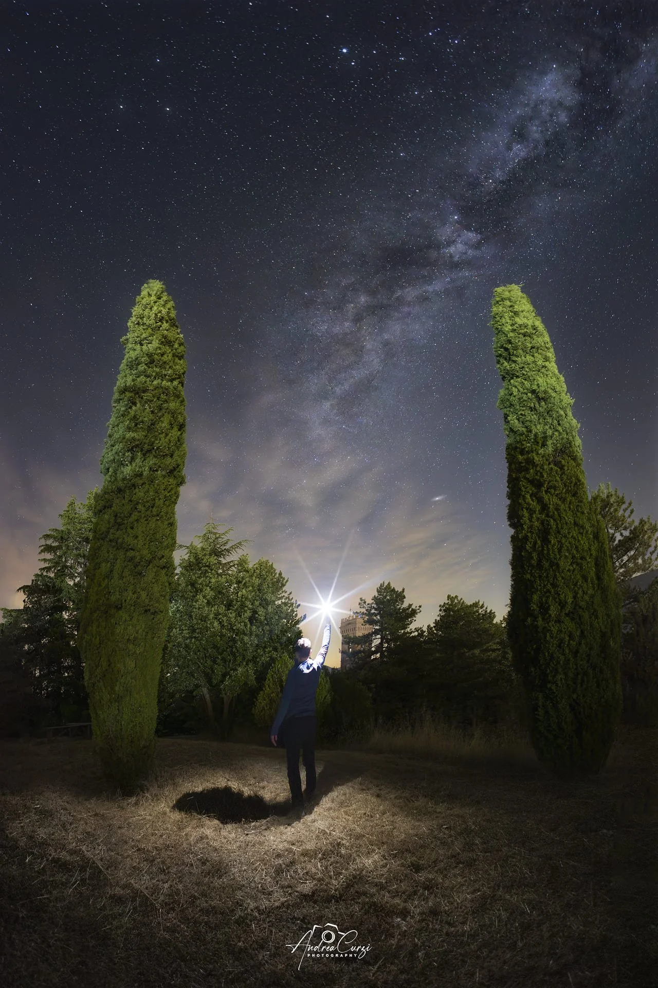 Persona che punta una torcia verso il cielo stellato e la coda della Via Lattea, tra due cipressi alti, di notte. Siamo sui Monti Sibillini, sul lago di Fiastra.
