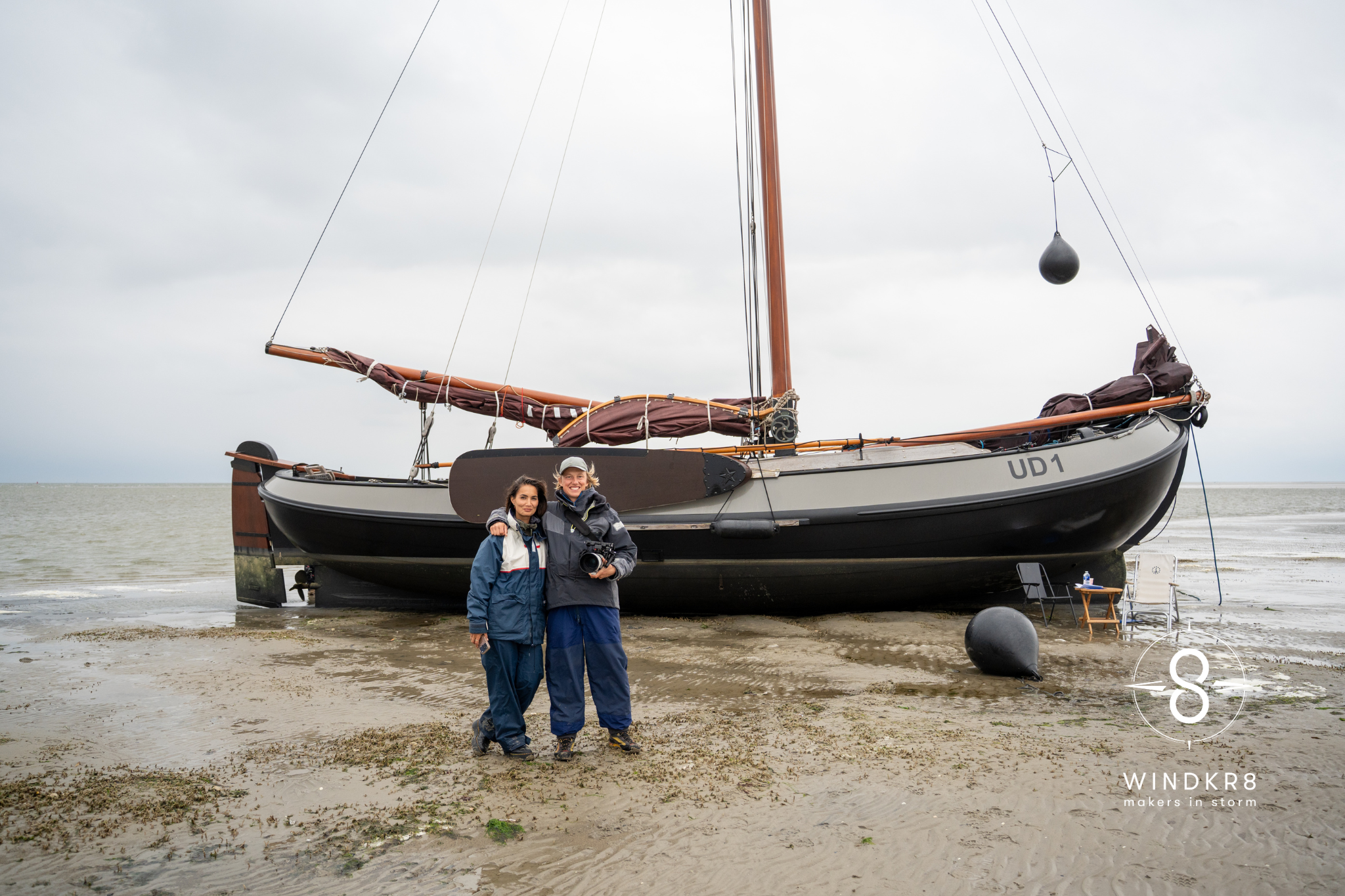 Twee mensen staan op het strand voor een grote zeilboot met de naam 'UD1'. Het is bewolkt en het lijkt stormachtig. Er staan ook enkele stoelen en een waterfles op het zand.