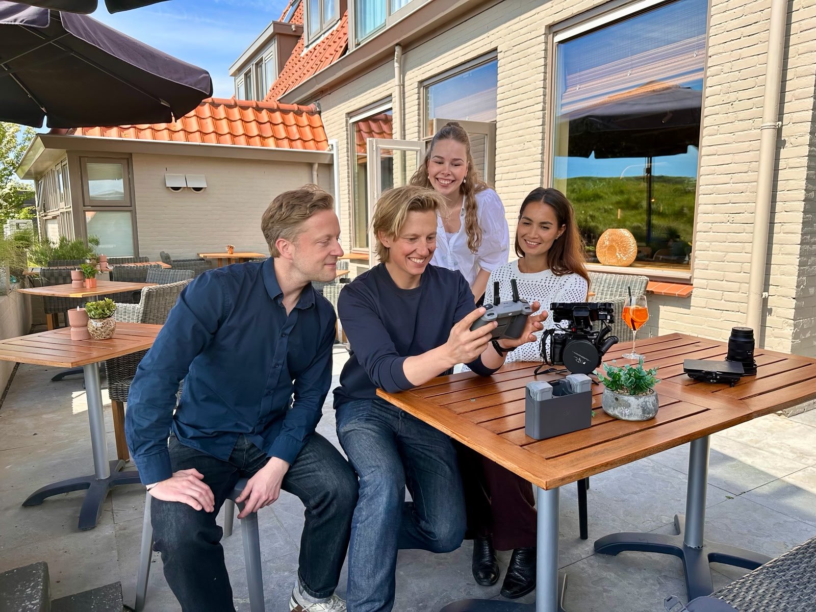 Vijf mensen die samen op een balkon zitten, met één persoon die een drone bestuurt, terwijl de anderen kijken en lachen. Er zijn tafels, planten en een drankje zichtbaar.