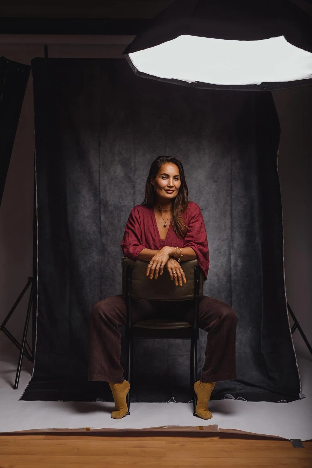 Een vrouw zit op een stoel voor een fotografiestudio, gekleed in een roze blouse en bruin broek, met een donkere achtergrond en een lampenbox boven haar.