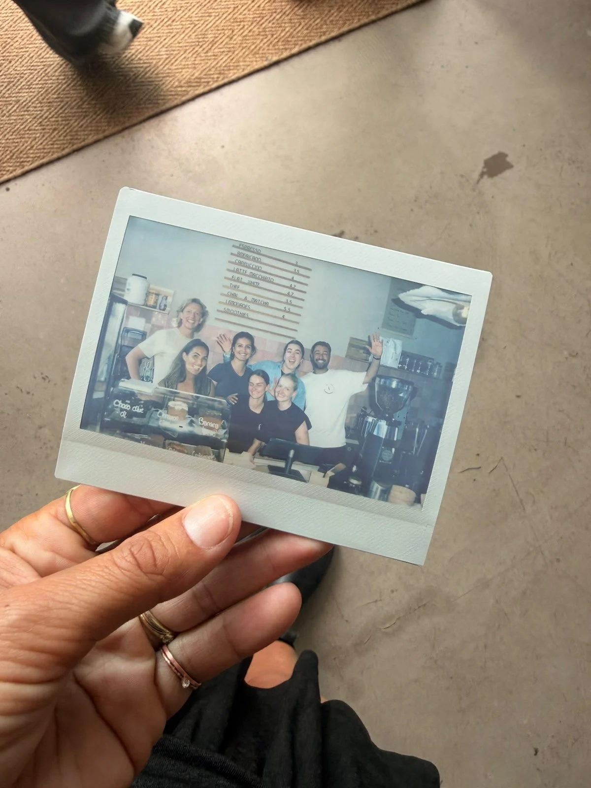 Een persoon houdt een polaroidfoto vast van zes mensen achter de counter van een koffiebar, waarvan vier vrouwen en twee mannen, die allemaal glimlachen en zwaaien.