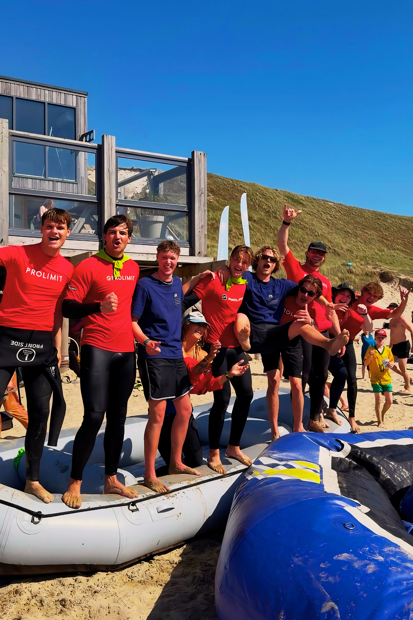 Groep mensen op een strand, klaar voor surfen, met surfplanken en een luchtband, onder een heldere blauwe lucht en dune achtergrond.