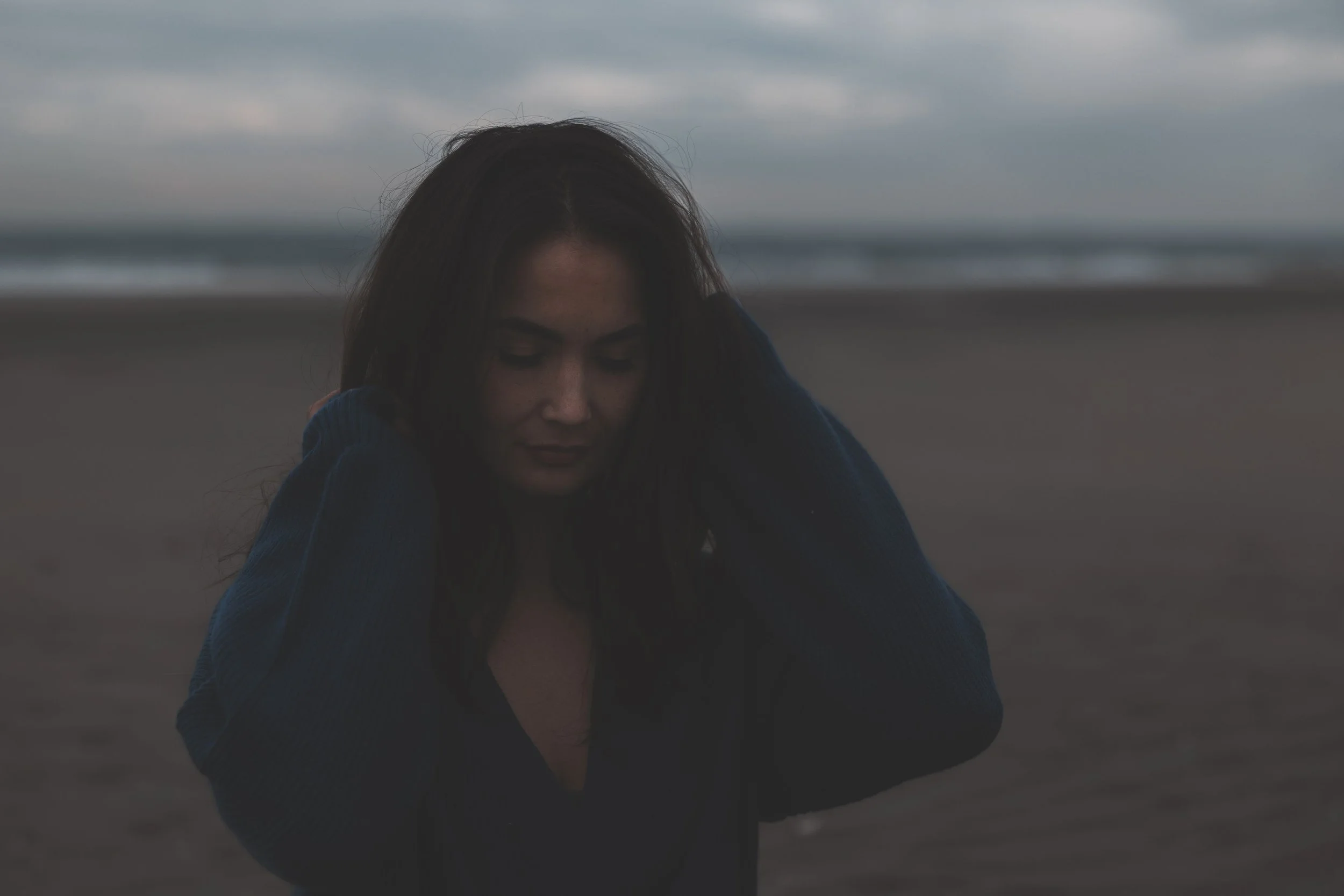 Een vrouw met lang donker haar op het strand, gekleed in een blauwe trui, met de zee en een bewolkte lucht op de achtergrond.