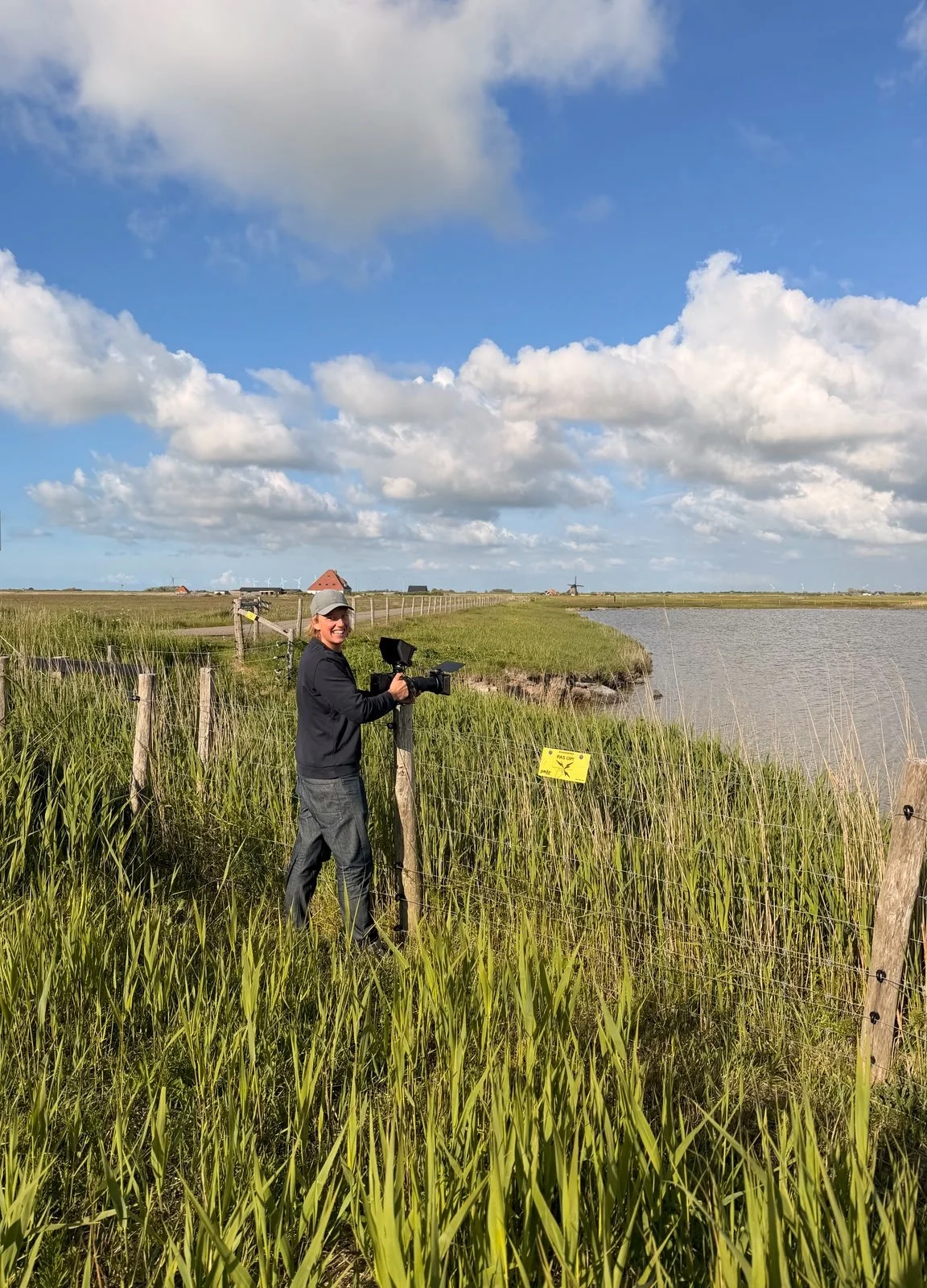 Een persoon met een videocamera bij een natuurrijke waterkant, omgeven door hoge groene grasmatten en een hek met een gele waarschuwing. Op de achtergrond zijn huizen, windmolens en een heldere blauwe lucht met witte wolken.