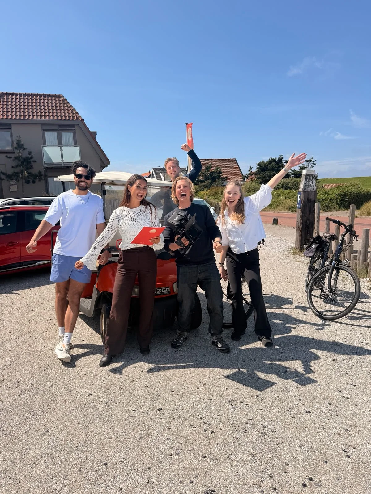 Vrouw en vier mensen vieren met een golfkar op een zonnige dag, enkele met camera's en documenten, naast fietsen.