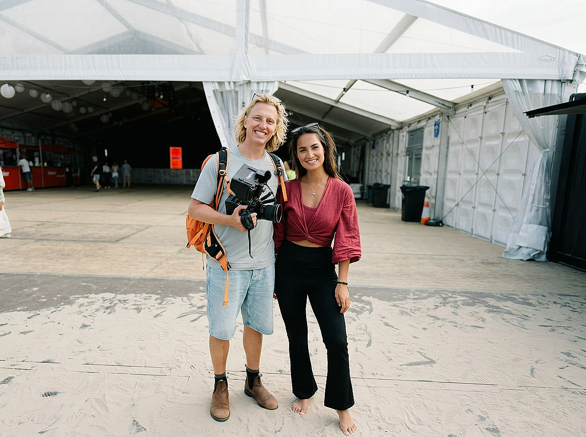 Twee vrouwen staan samen onder een grote witte tent, lachen en poseren voor de camera. Eén vrouw heeft blond haar, draagt een grijs T-shirt, korte broek en laarzen, de andere vrouw heeft donker haar en draagt een rode top en zwarte broek.