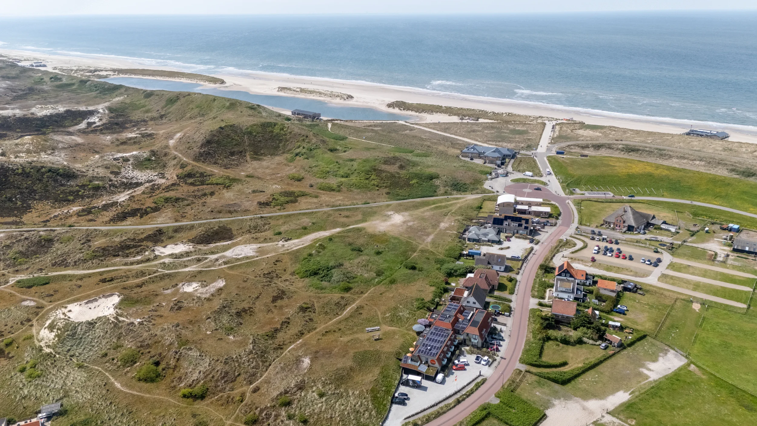 Luchtfoto van een kustgebied met zandstranden, heuvels en een klein dorp met huizen en wegen. Aan de achtergrond de oceaan met golven.