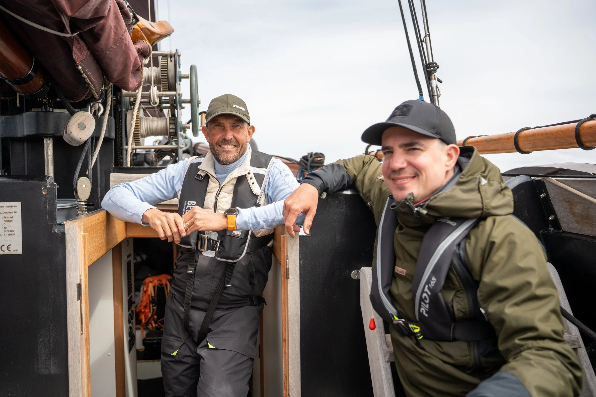 Twee mannen in outdoor kleding en petten, lachen en poseerend op een schip, met technische apparatuur en touwen op de achtergrond.